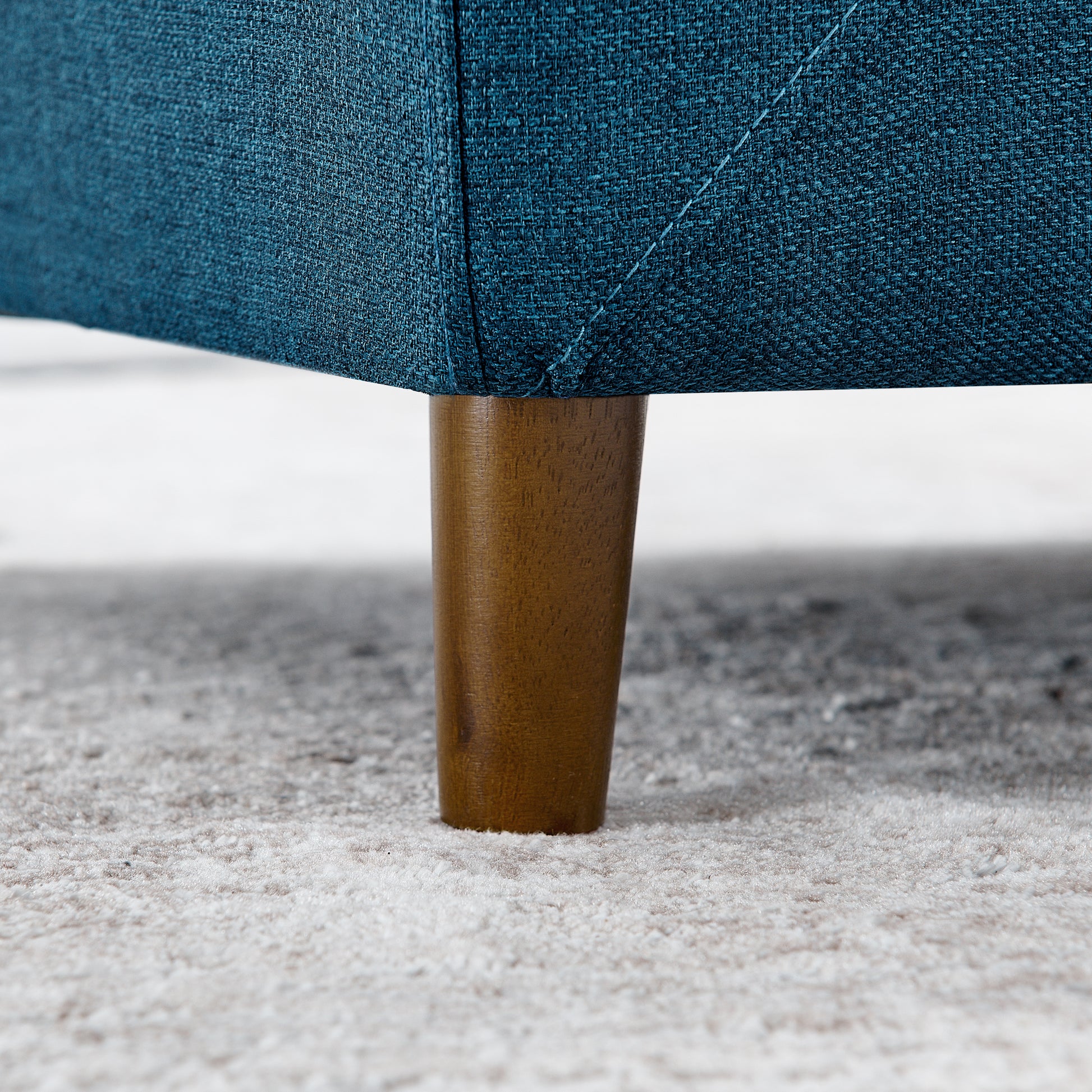 Closeup of a teal textured fabric sofa leg with a tapered wooden base on a neutral rug