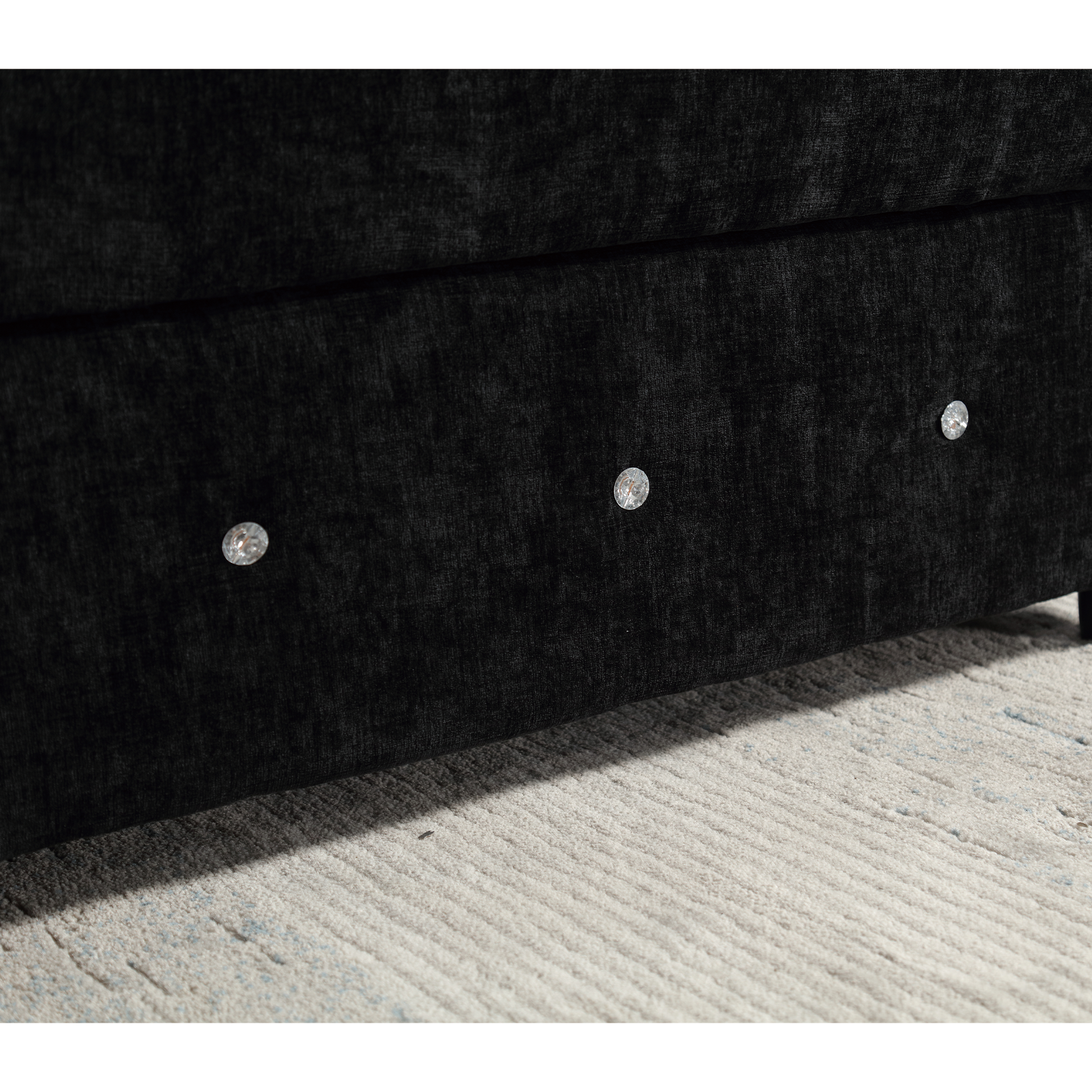 Closeup of a black velvet divan bed base with crystal button tufting on a light rug