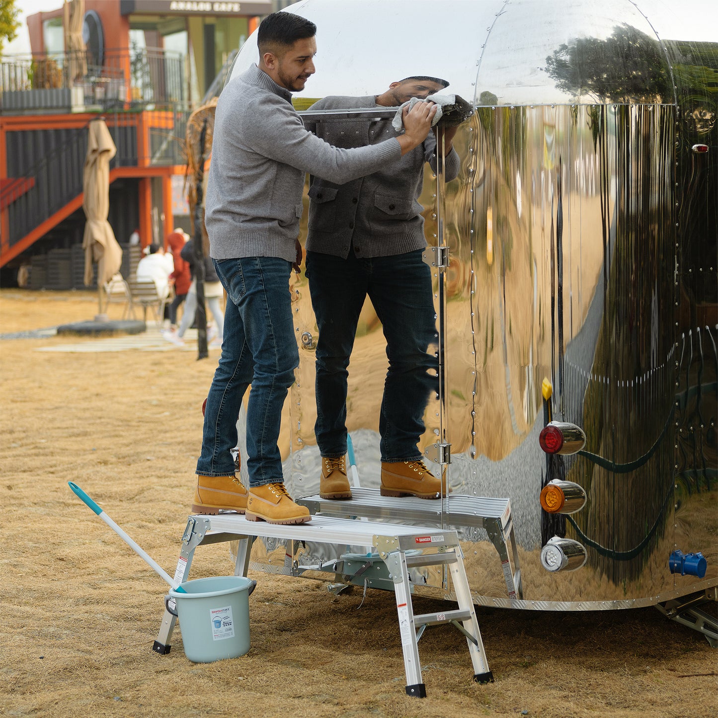 Men cleaning shiny Airstream trailer with cloth on aluminum platform ladder