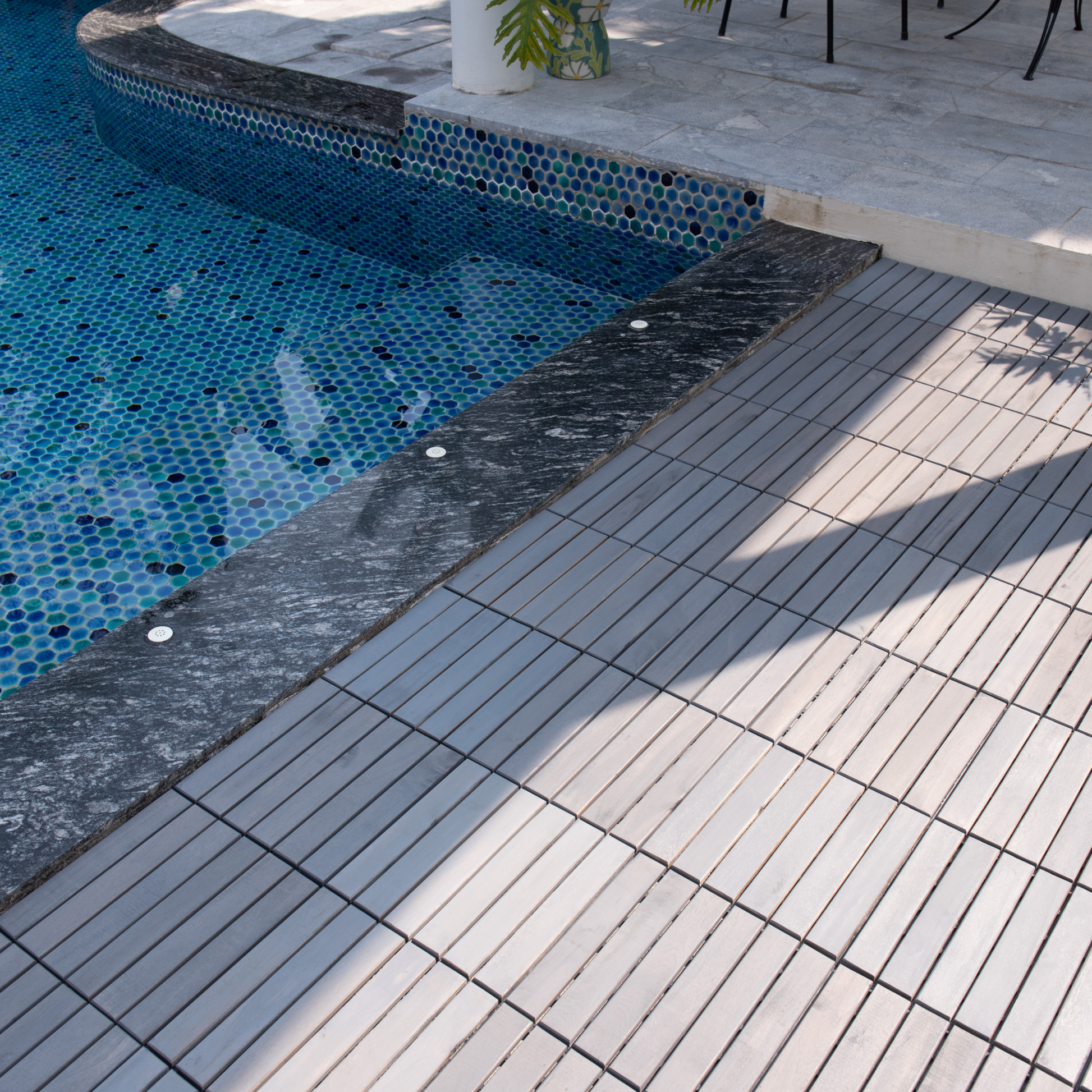 Blue mosaic tiles form the pool wall transitioning to a dark granite edge and light gray deck tiles