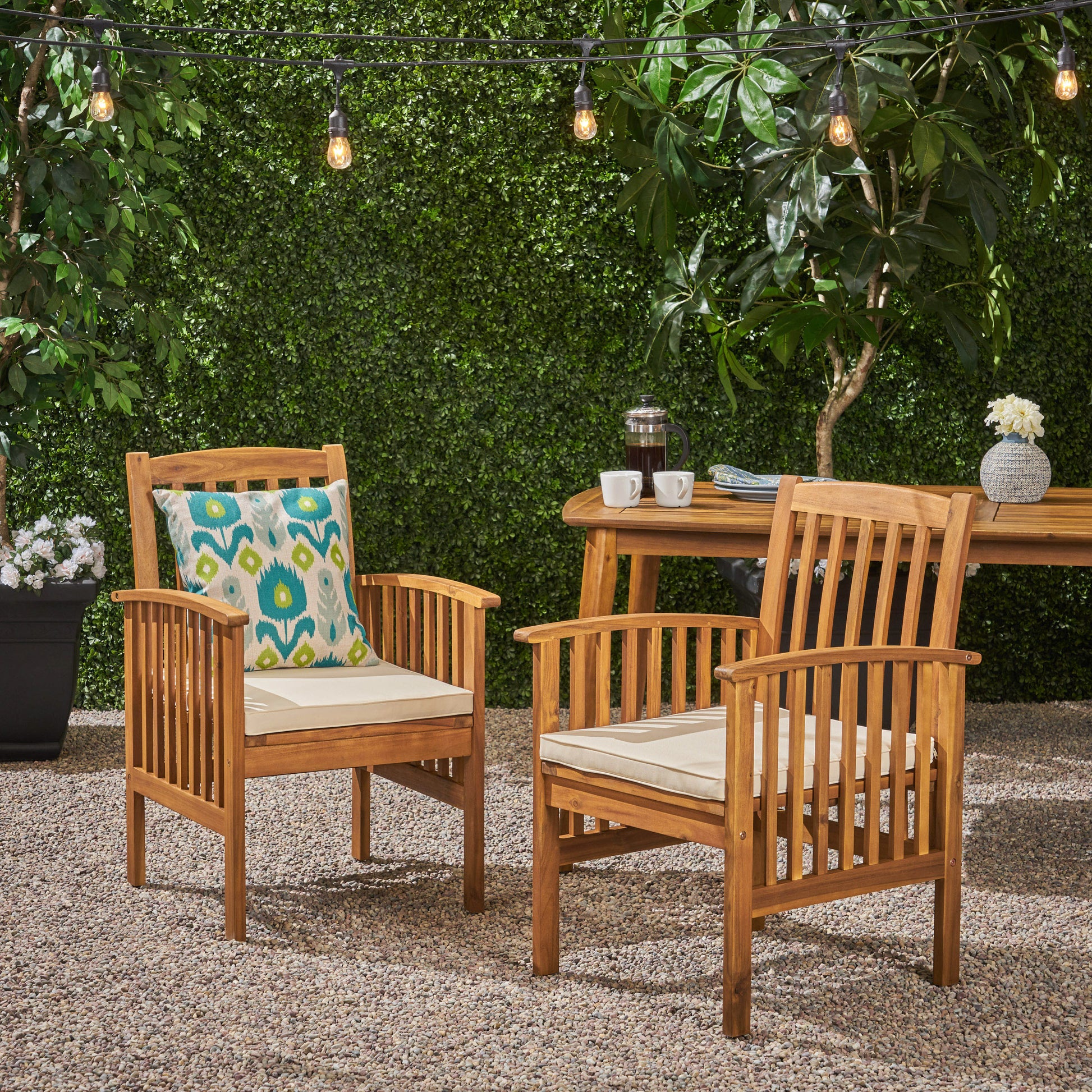 Two warmtoned wooden patio chairs with cream cushions one featuring a teal and green patterned throw pillow