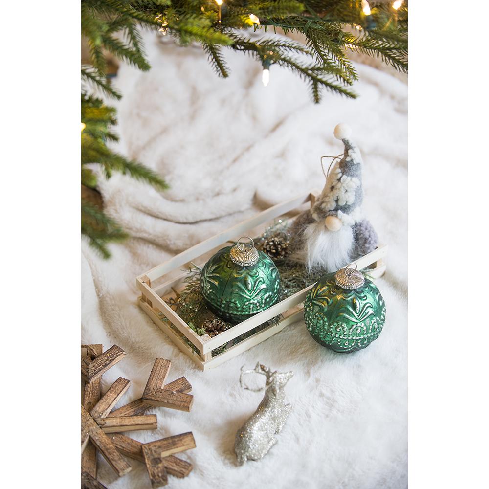 Two emerald green glass Christmas ornaments with embossed white floral patterns sit in a wooden crate