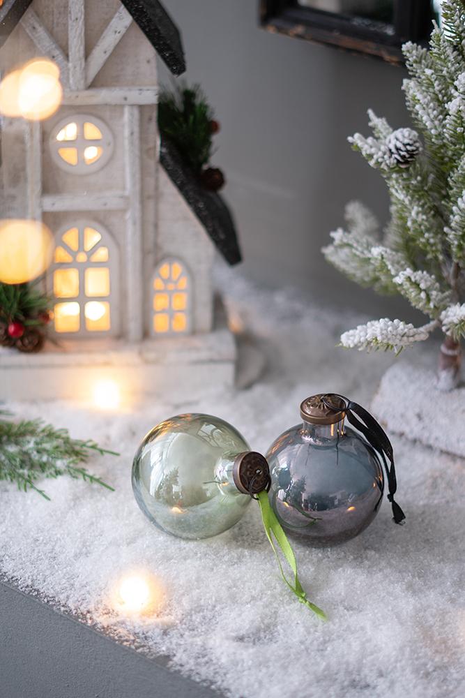 Two vintagestyle glass Christmas ornaments one pale green one metallic gray resting on faux snow
