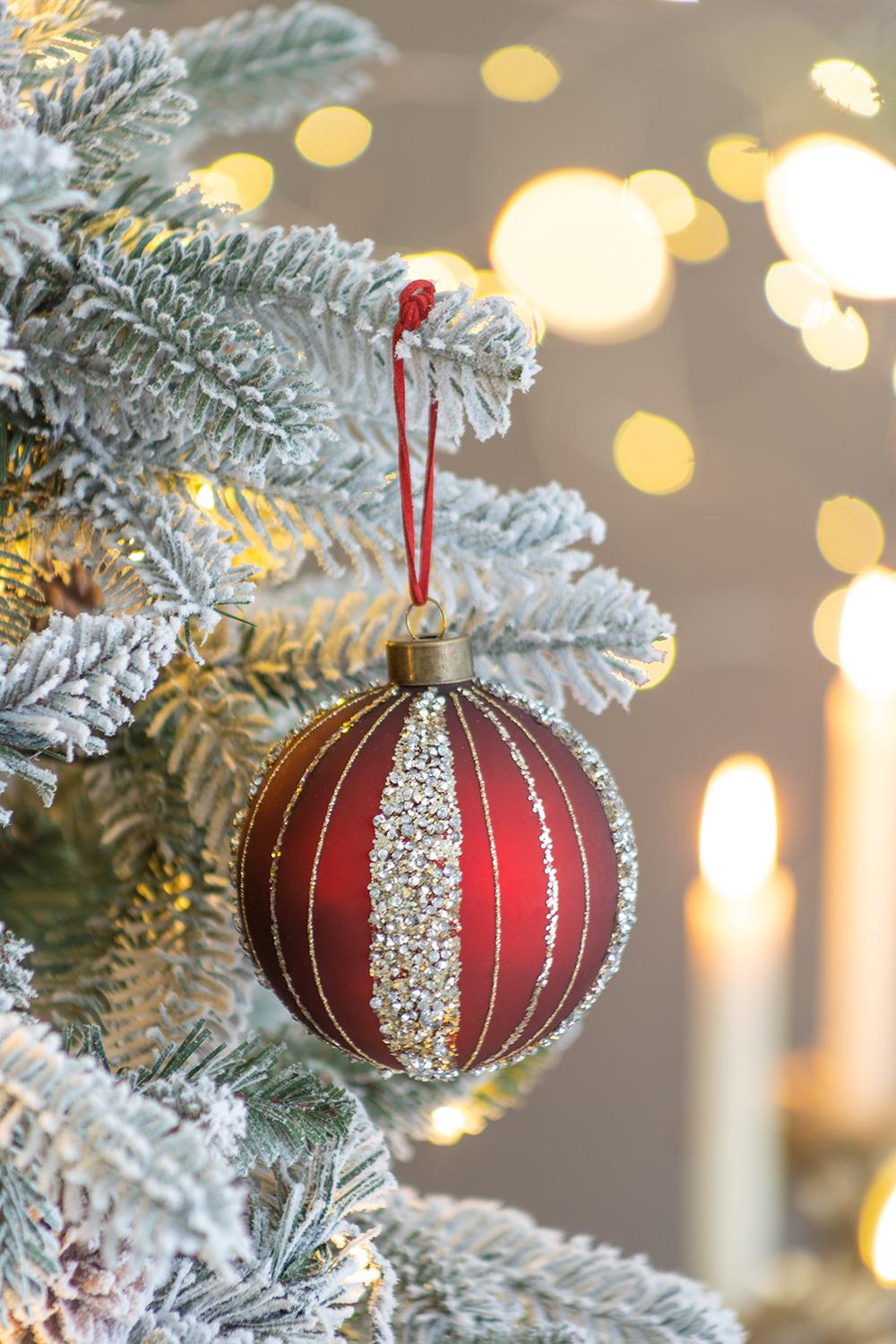 Red glass Christmas bauble with glitter stripes and gold accents hanging on frosted tree branch
