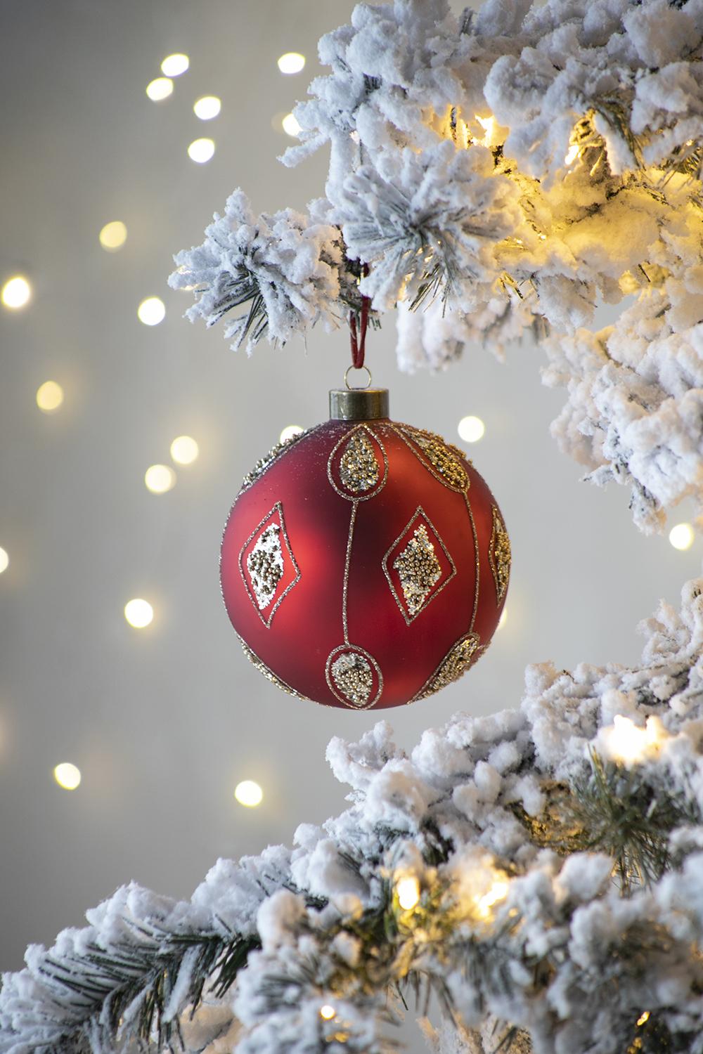 Red glass Christmas ornament with gold diamond pattern and glitter accents hanging on a flocked tree branch