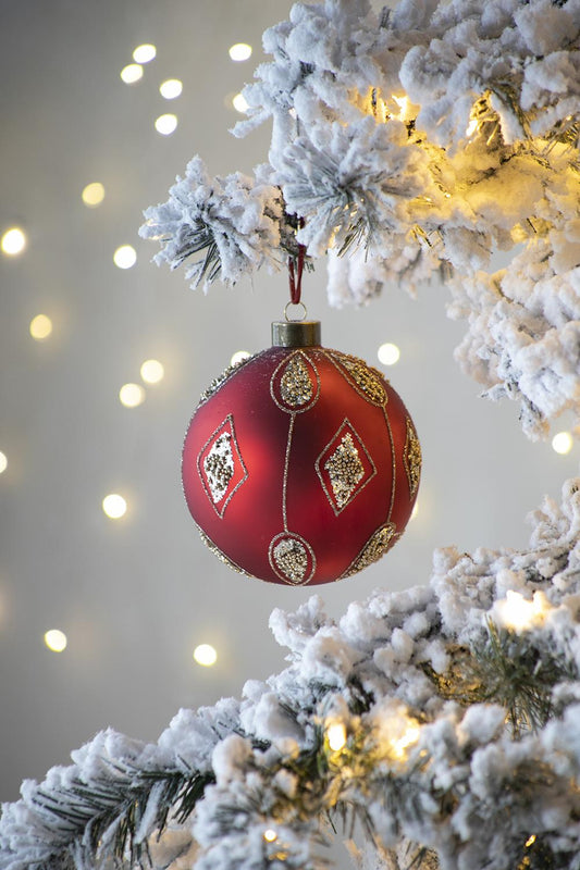Red glass Christmas ornament with gold diamond pattern and glitter accents hanging on a flocked tree branch