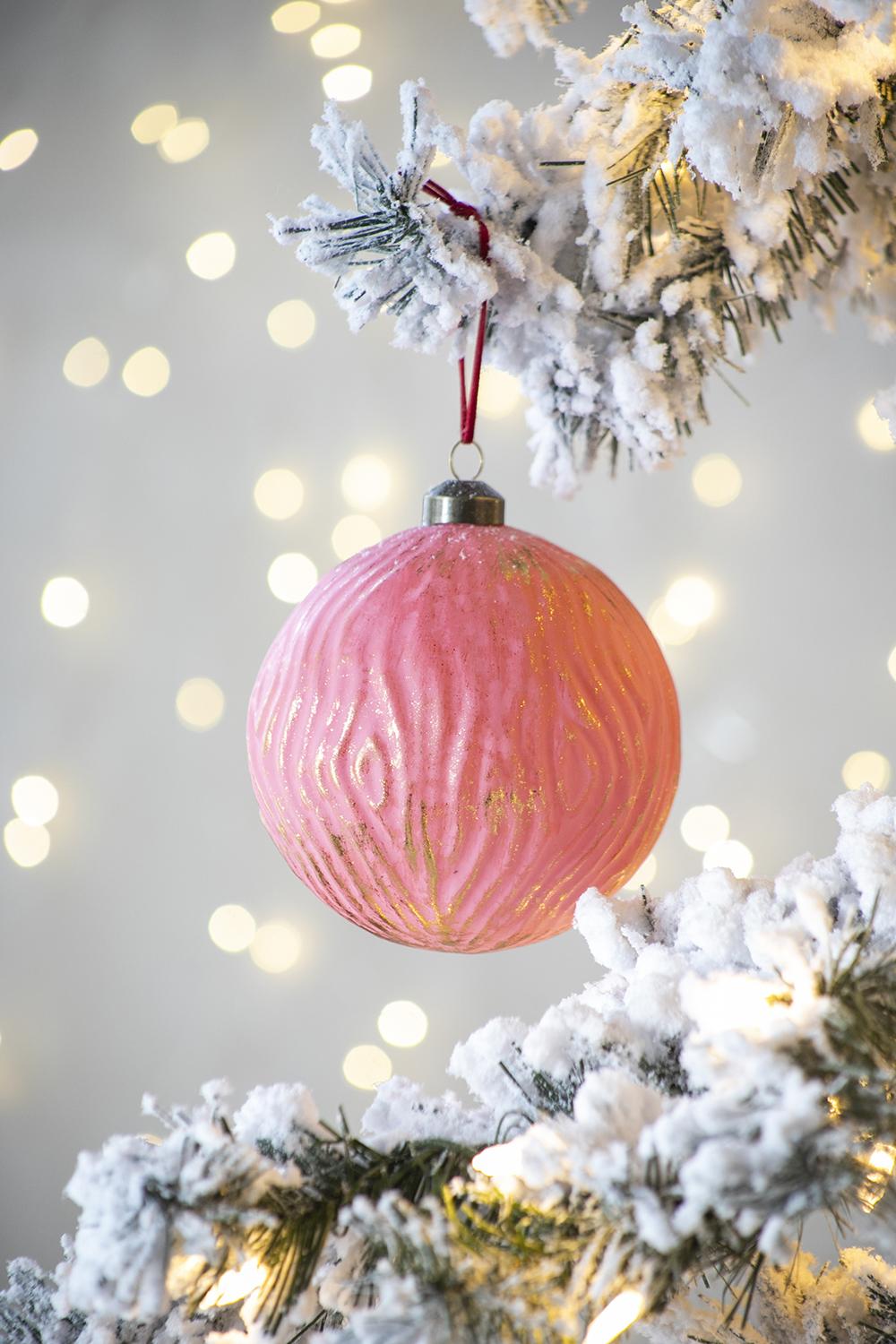 Pink glass Christmas ornament with gold swirling texture hanging on a flocked tree branch