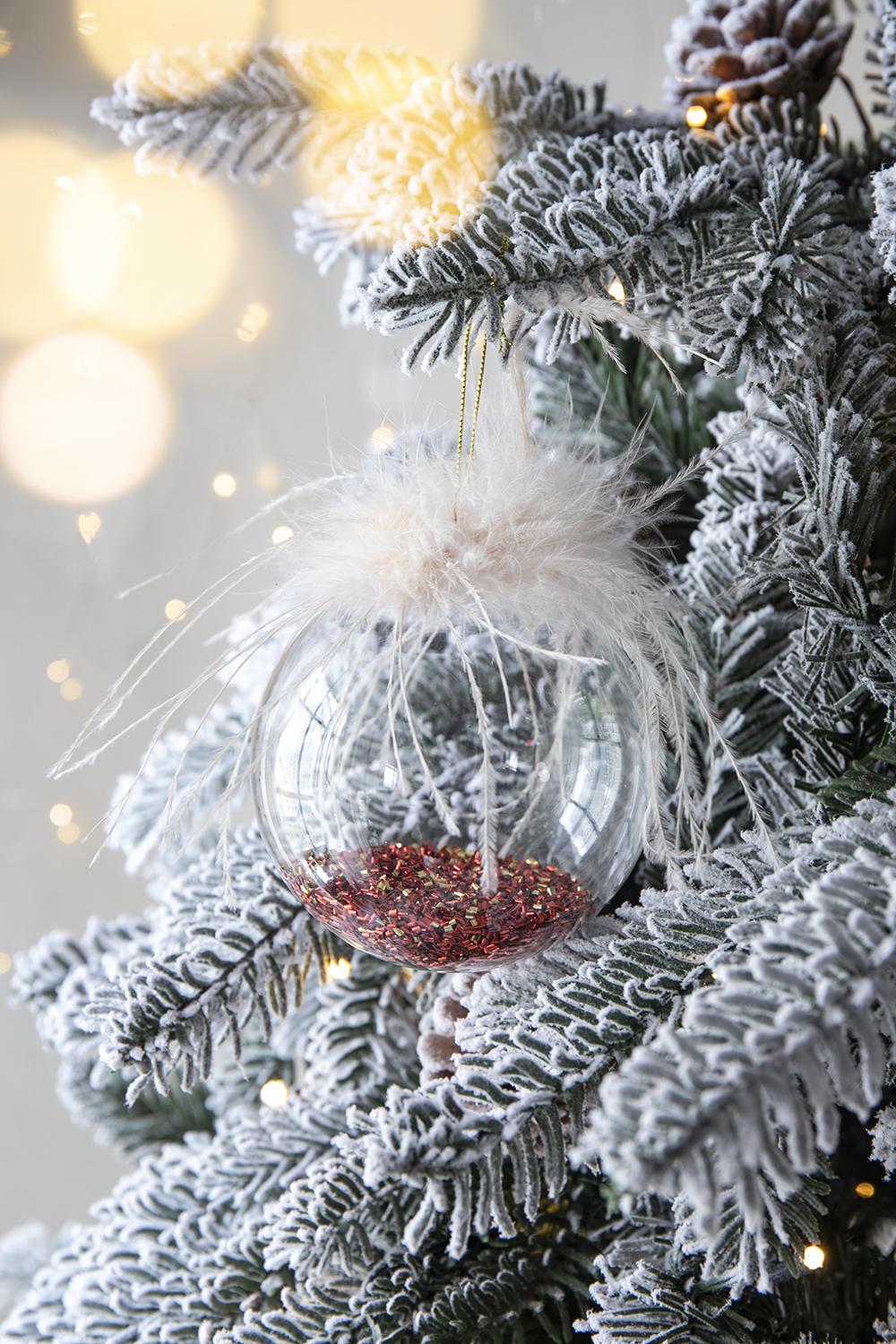 Clear glass Christmas ornament with tan feathers and red glitter inside hanging on a frosted tree branch