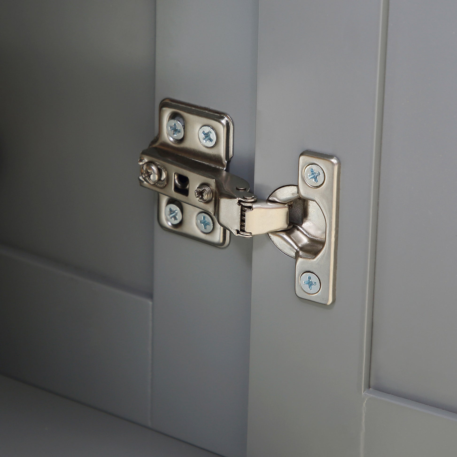 Closeup of a brushed nickel cabinet hinge on a grey wooden door panel showing screw details