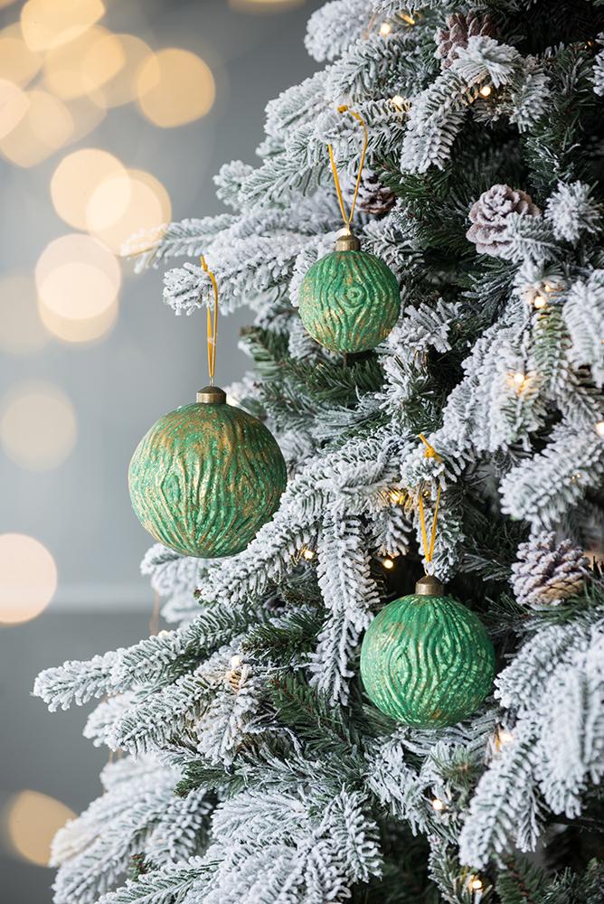 Three green textured christmas ornaments with gold accents hang on a flocked tree