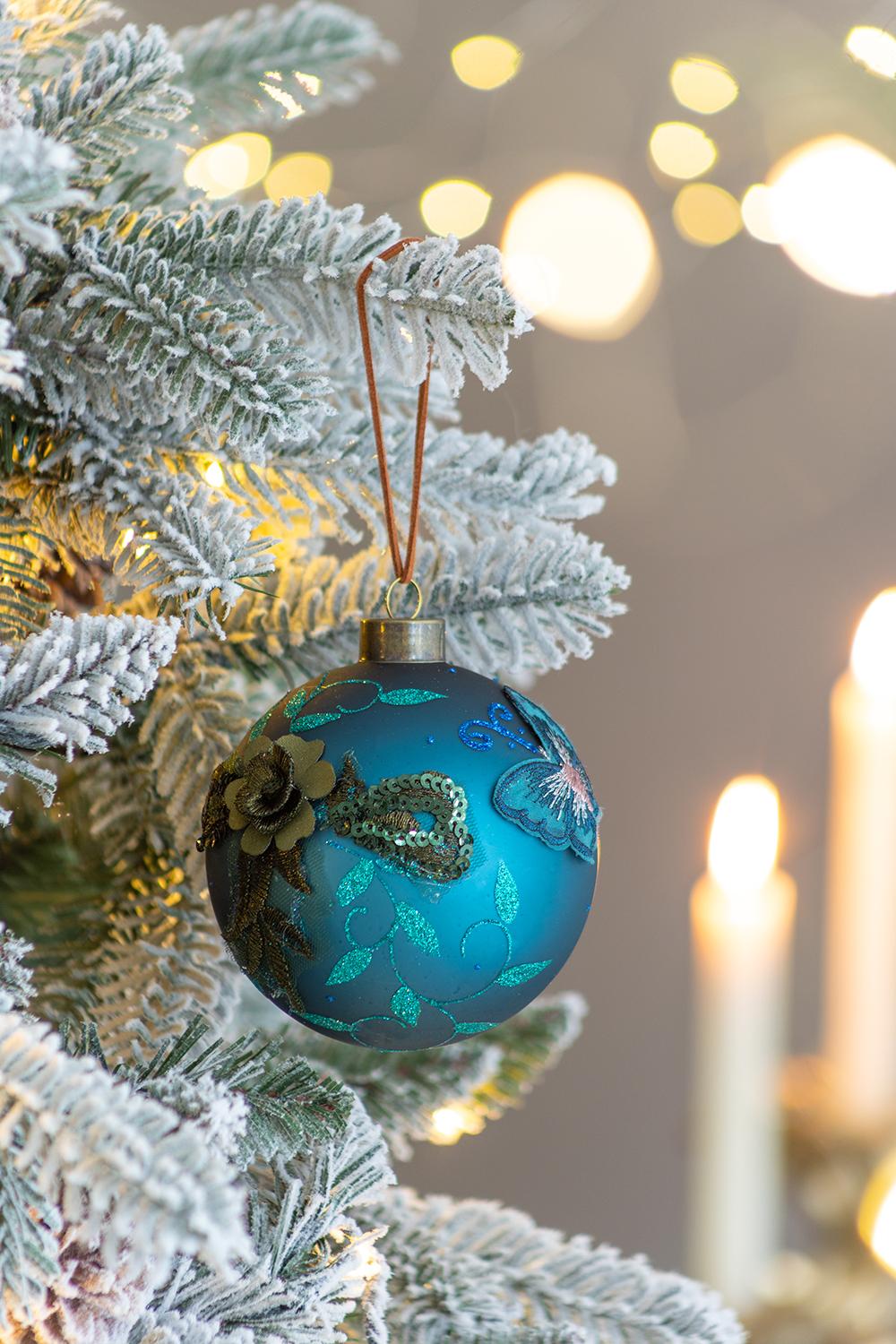 Teal glass Christmas ornament with embroidered flowers and sequin details on a frosted tree branch