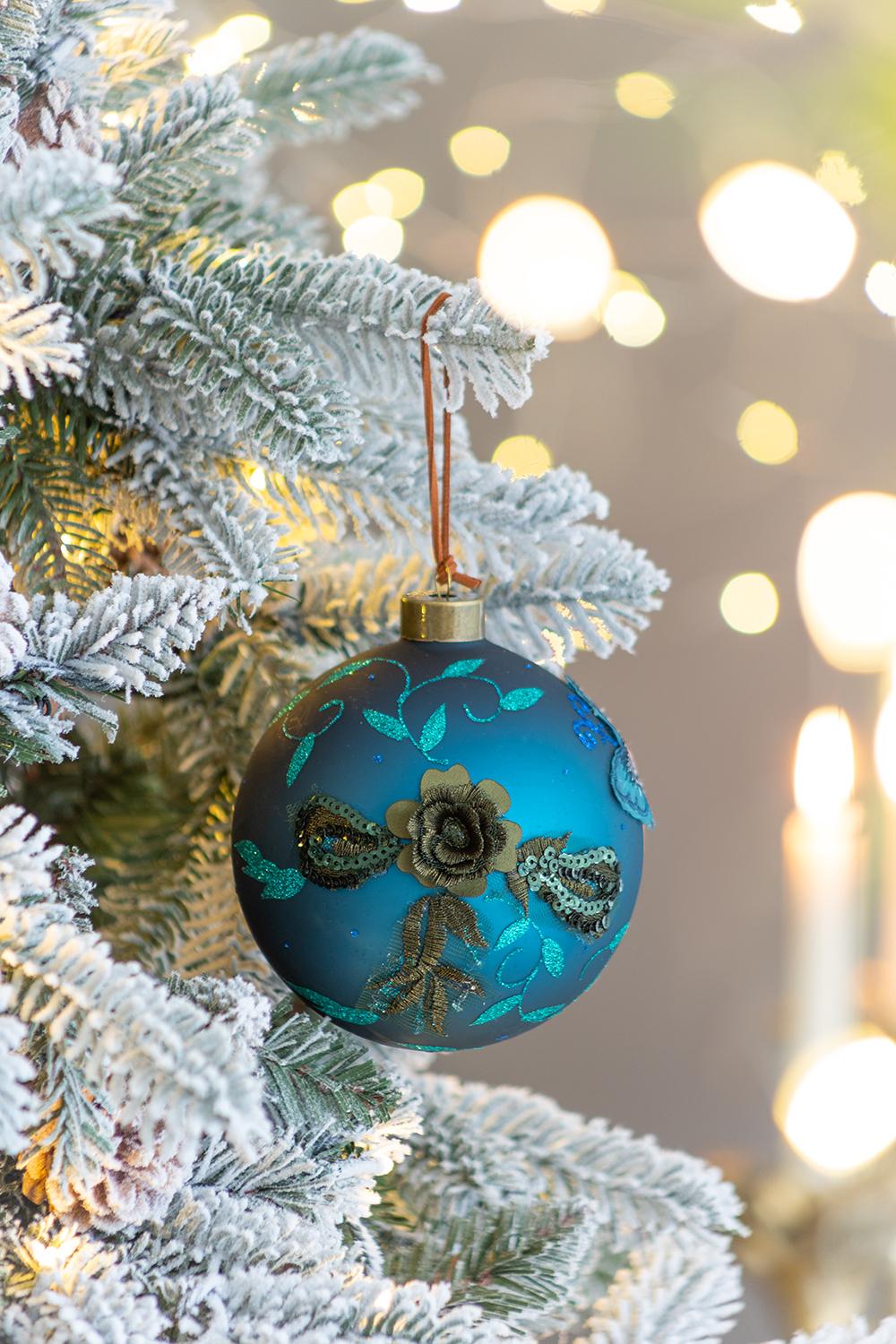 Teal glass Christmas ornament with embroidered floral and sequin details on a flocked tree
