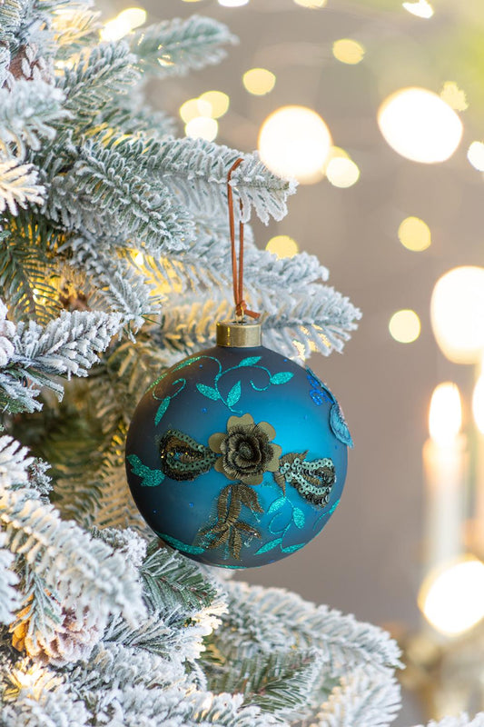 Teal glass Christmas ornament with embroidered floral and sequin details on a flocked tree