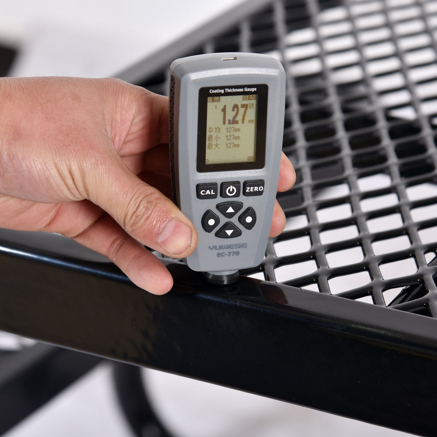 Hand holding a grey coating thickness gauge measuring a black metal grille