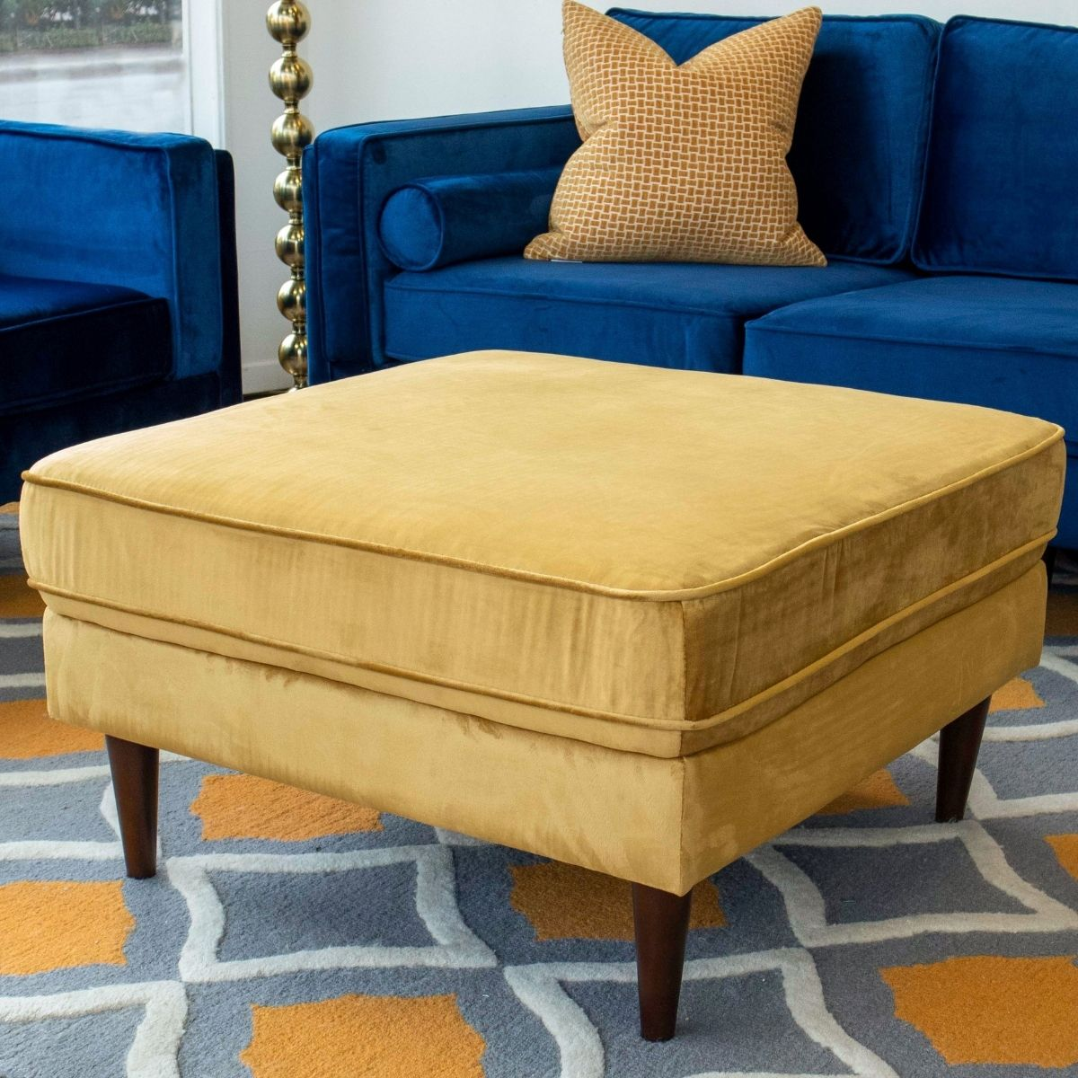 Gold velvet upholstered ottoman with plush cushioning featuring dark wooden legs for modern living room decor