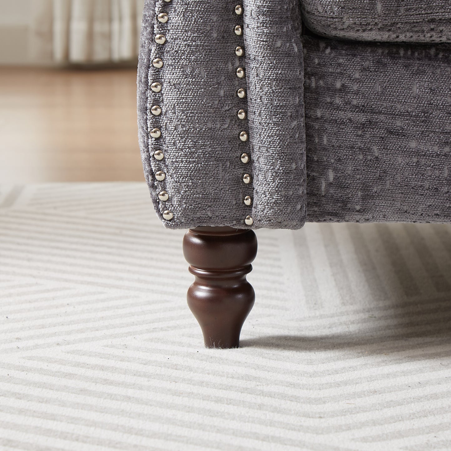 Closeup of a gray upholstered armchair with nailhead trim and a carved wooden leg on a striped rug