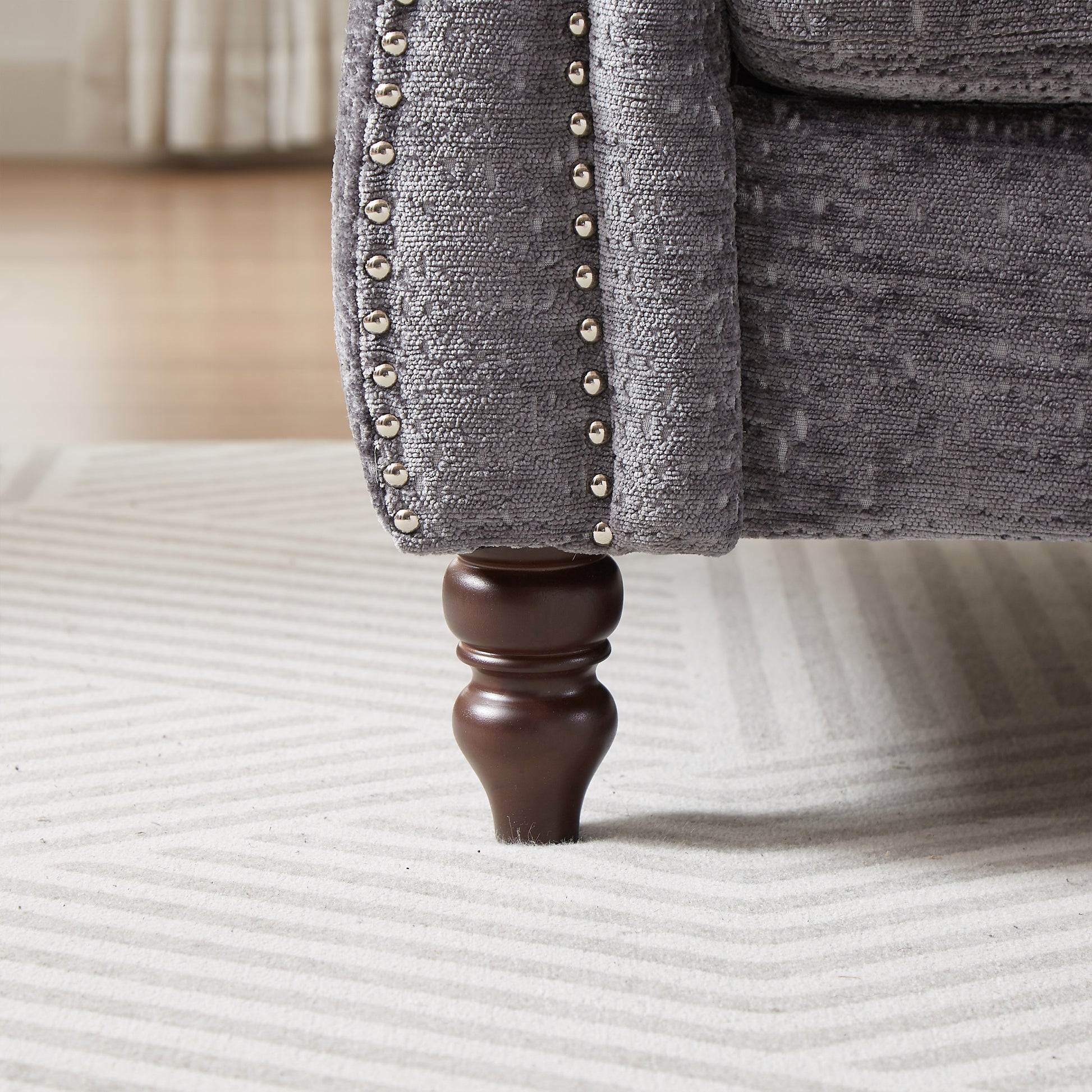 Closeup of a gray upholstered armchair with nailhead trim and a carved wooden leg on a striped rug