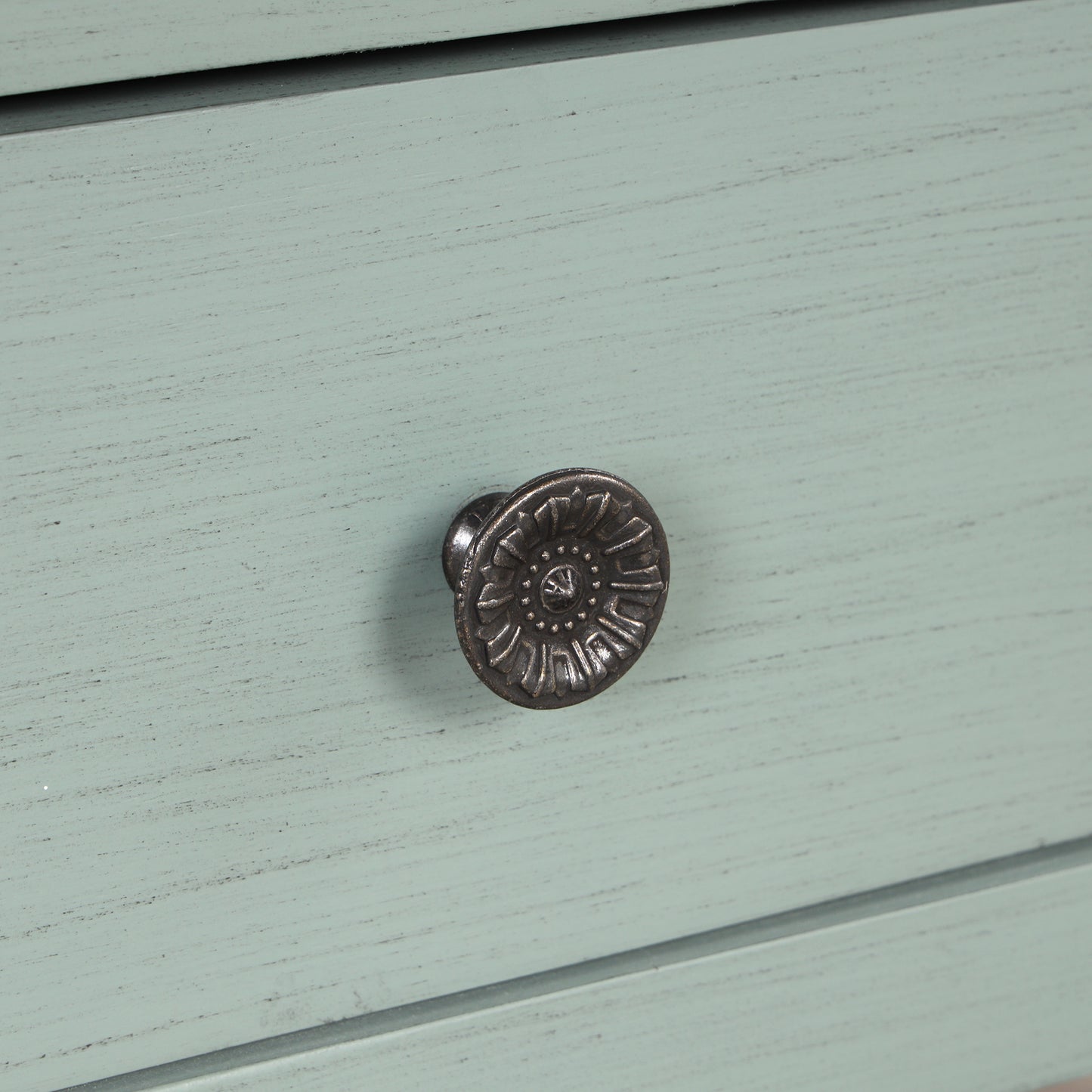 Closeup of a vintagestyle round antique brass drawer pull with floral detailing on a pale green distressed wood dresser