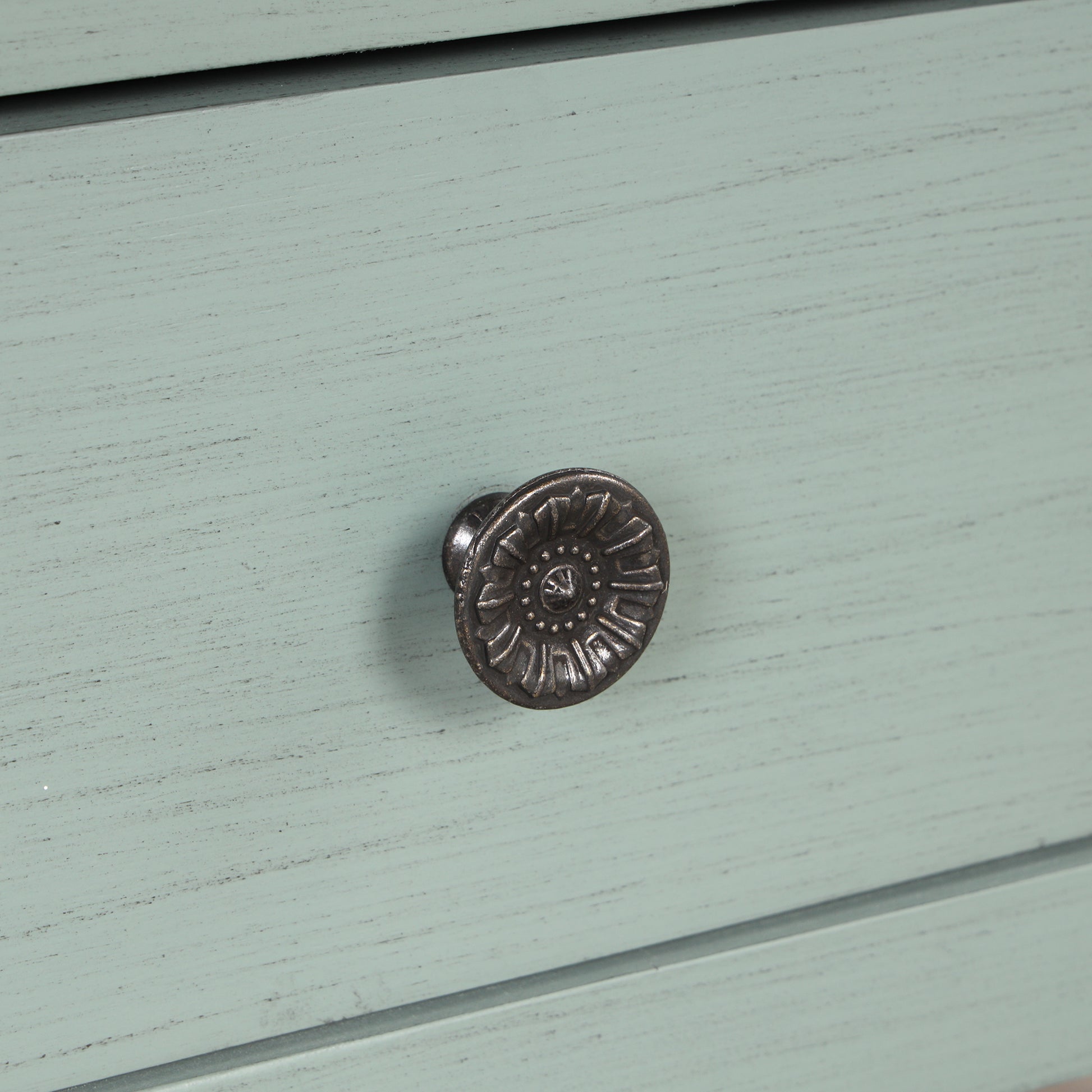 Closeup of a vintagestyle round antique brass drawer pull with floral detailing on a pale green distressed wood dresser