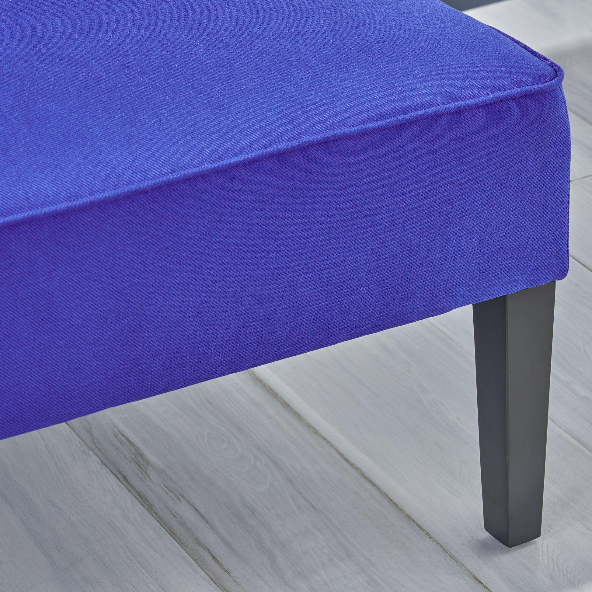 Close up of a rich royal blue upholstered ottoman with piped edges and dark tapered legs