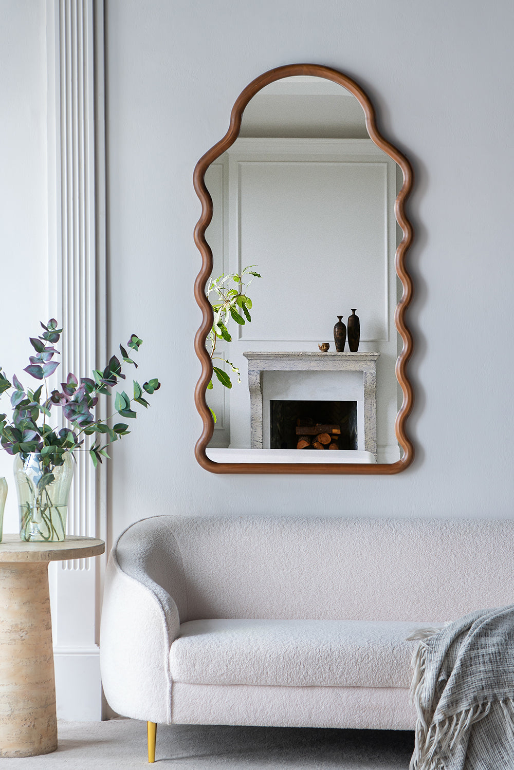 Curved wooden frame mirror with wavy edges reflecting a fireplace mantel and vases