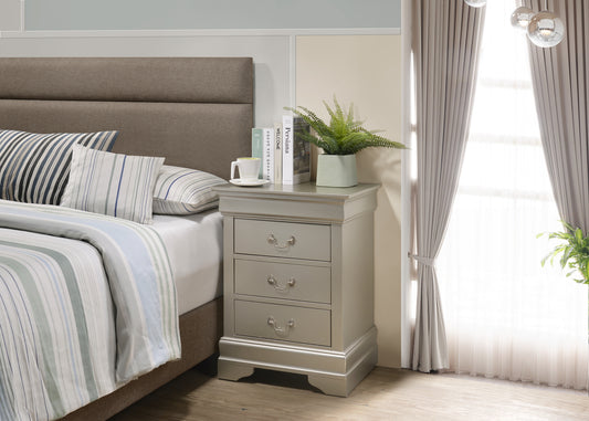 Silver finished nightstand with three drawers and decorative hardware beside a bed