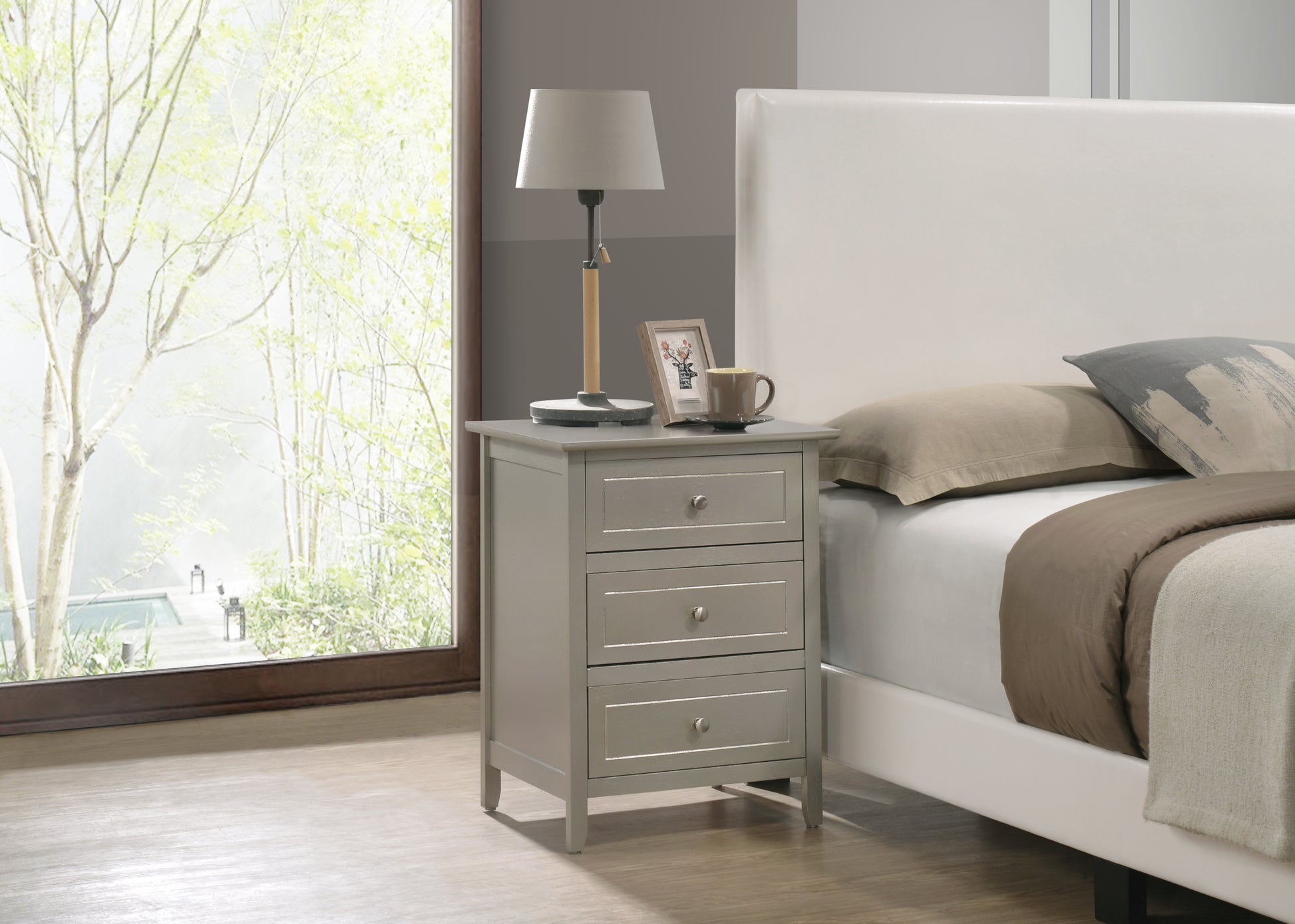 Gray painted wood bedside table with three drawers and silver hardware next to a white upholstered bed
