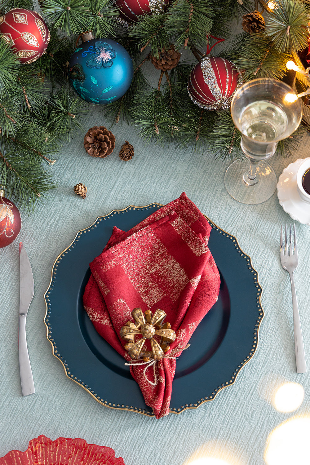 Dark teal charger plate with gold beaded rim holding a red goldpatterned napkin with gold brooch