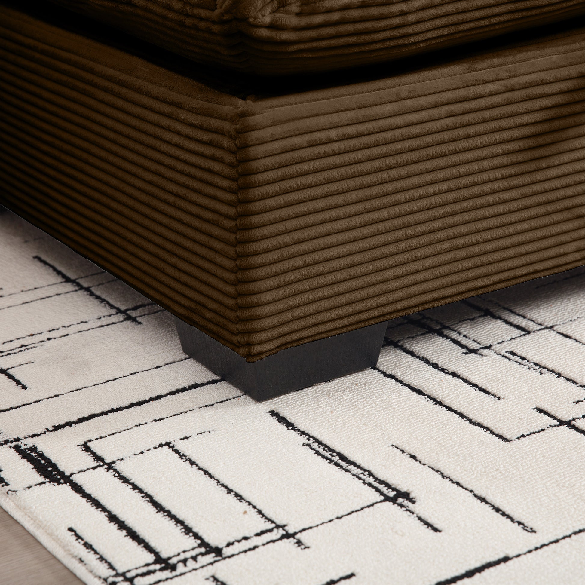 Closeup of a plush brown corduroy ottoman corner with black wooden legs on a cream rug
