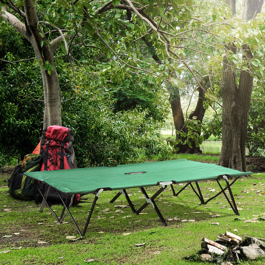 Green camping cot with metal frame set up on grass near trees and backpacks