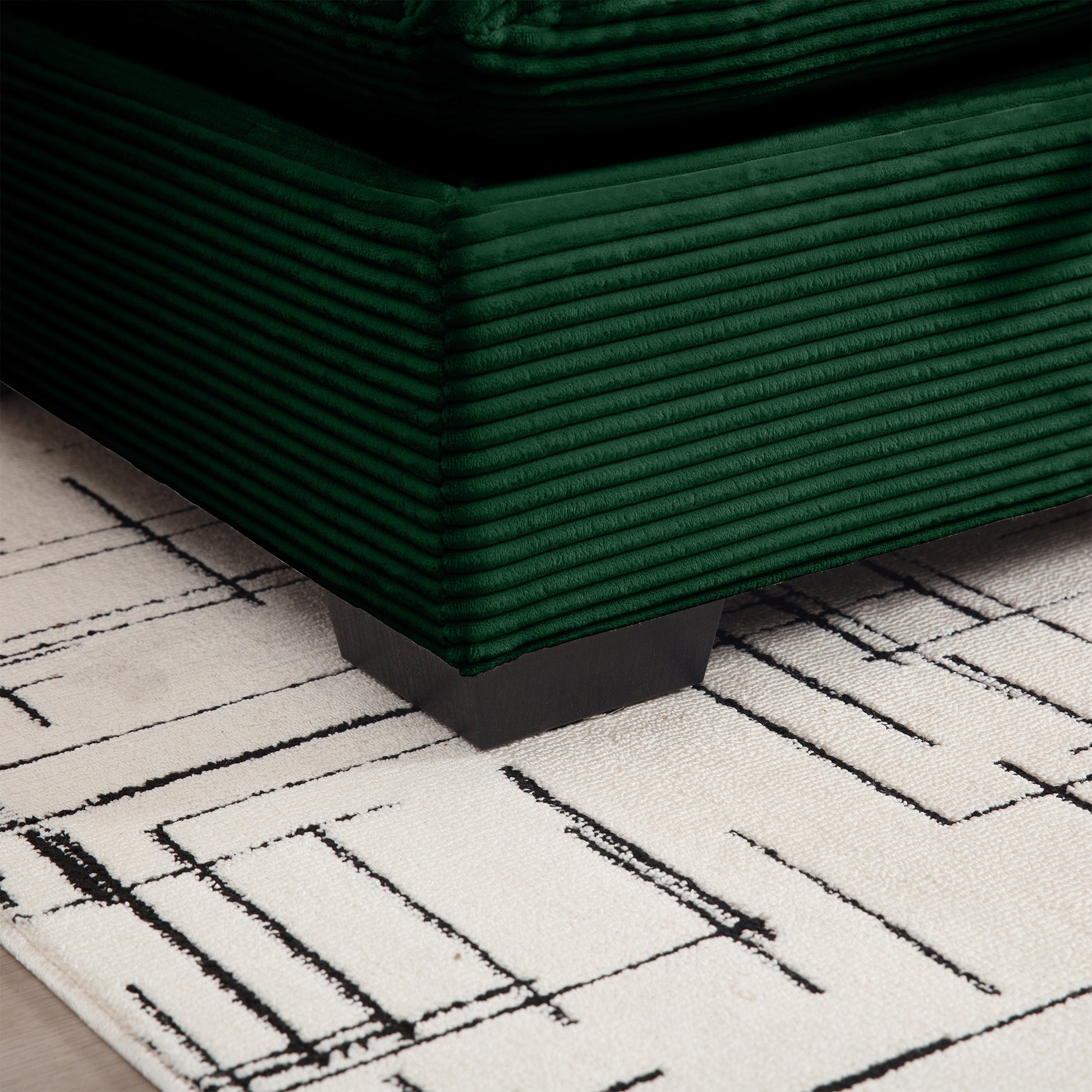 Closeup of a deep emerald green corduroy sofa cushion with a black wooden leg resting on a cream rug with black abstract lines