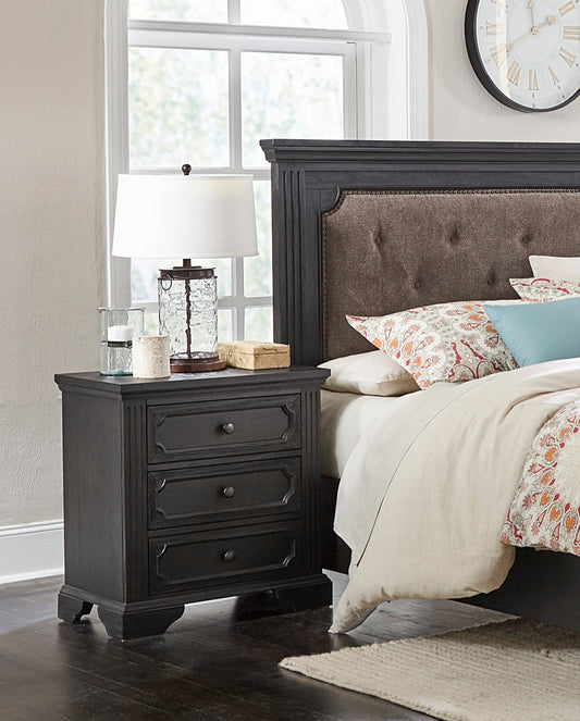 Dark wood threedrawer nightstand with carved detailing beside upholstered headboard