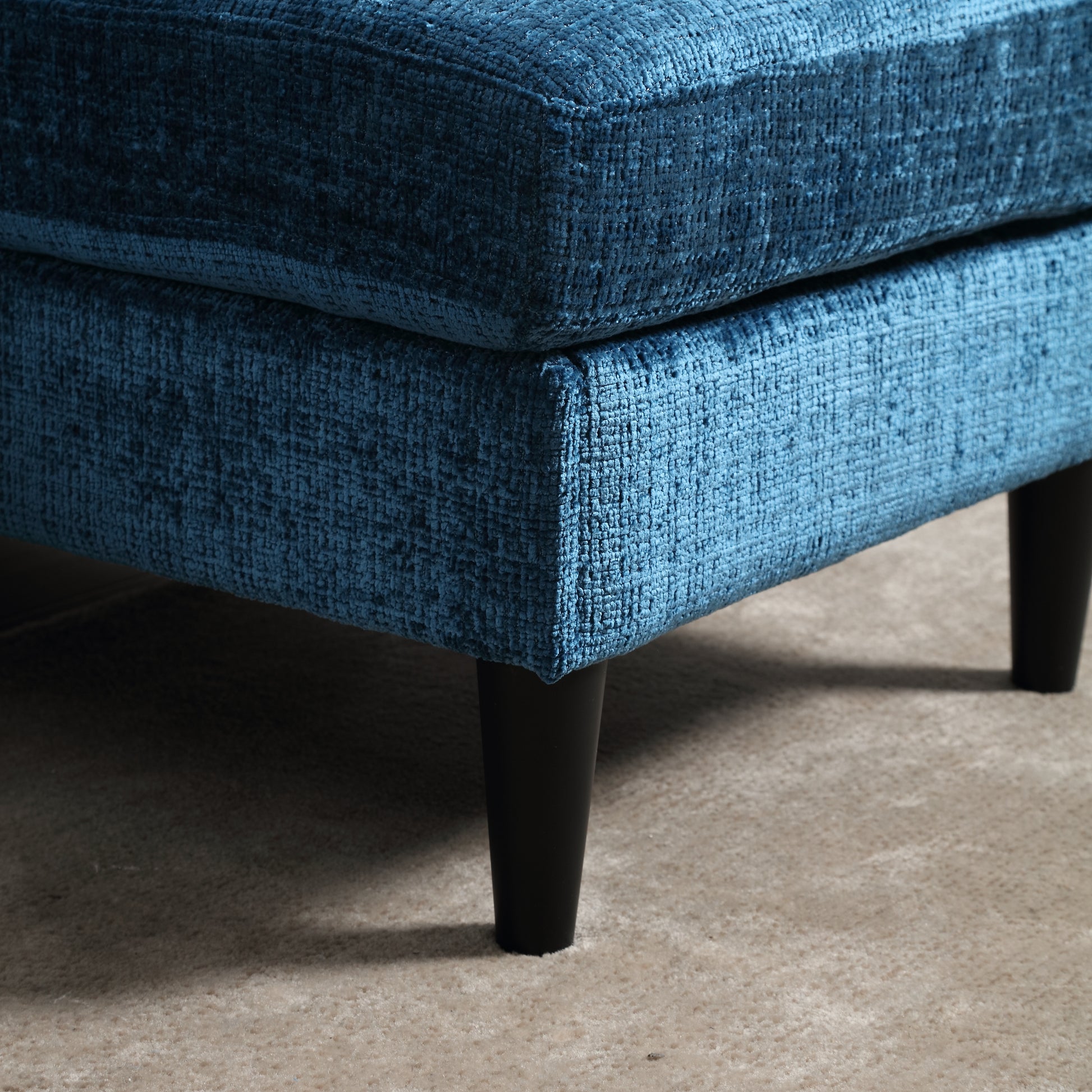 Closeup of a plush teal velvet ottoman corner with dark tapered legs on a textured rug