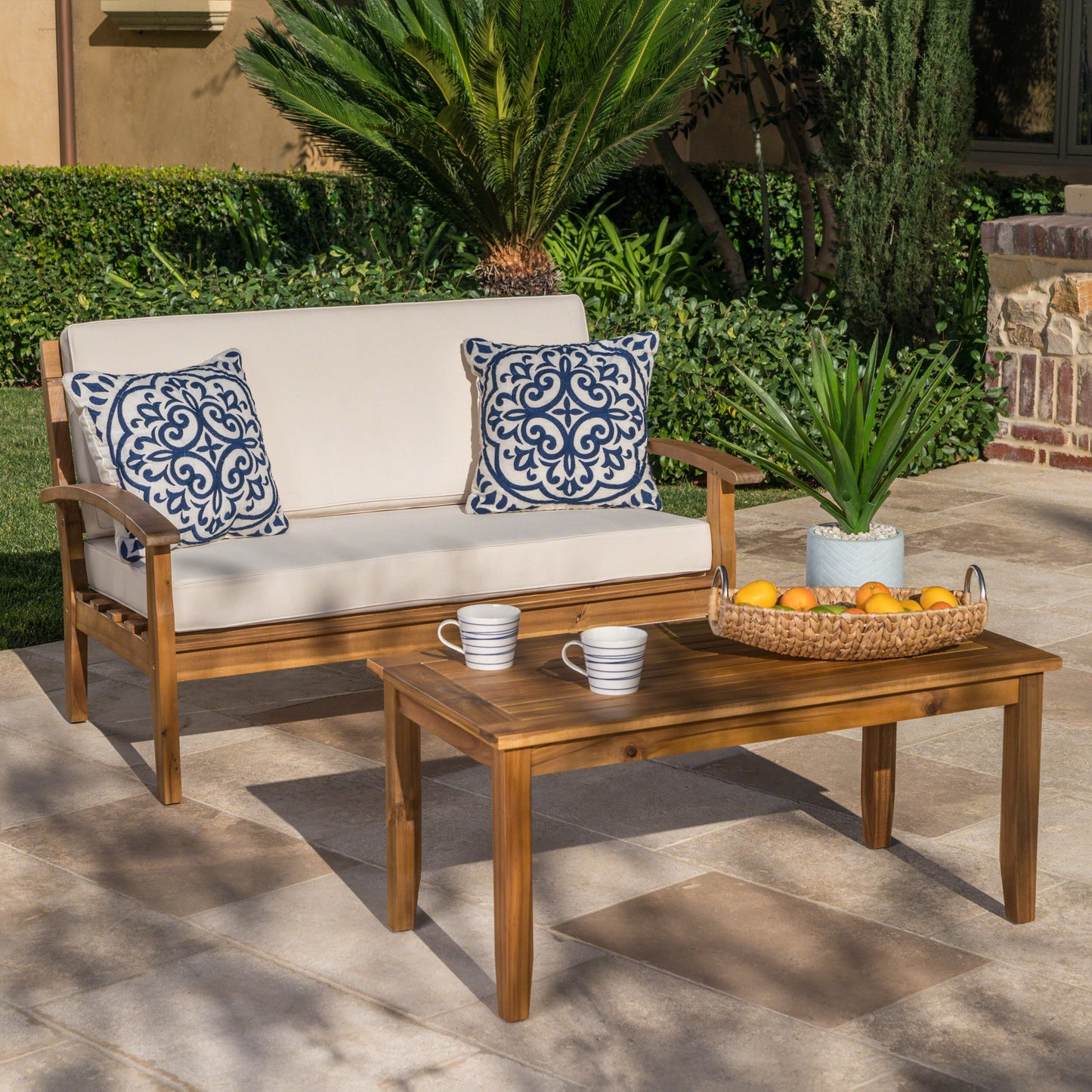 Acacia wood patio loveseat with plush beige cushions and blue patterned outdoor pillows