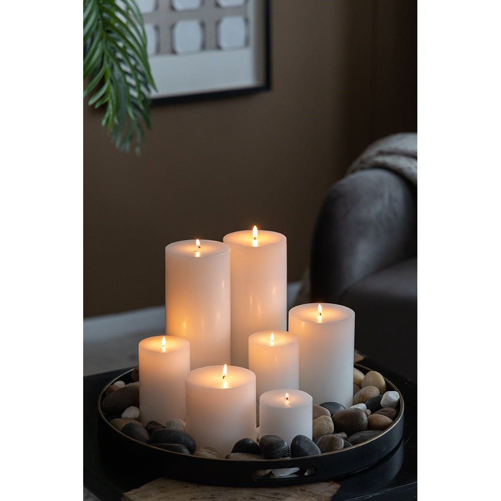 Group of six white pillar candles with flickering flames on a black tray with stones