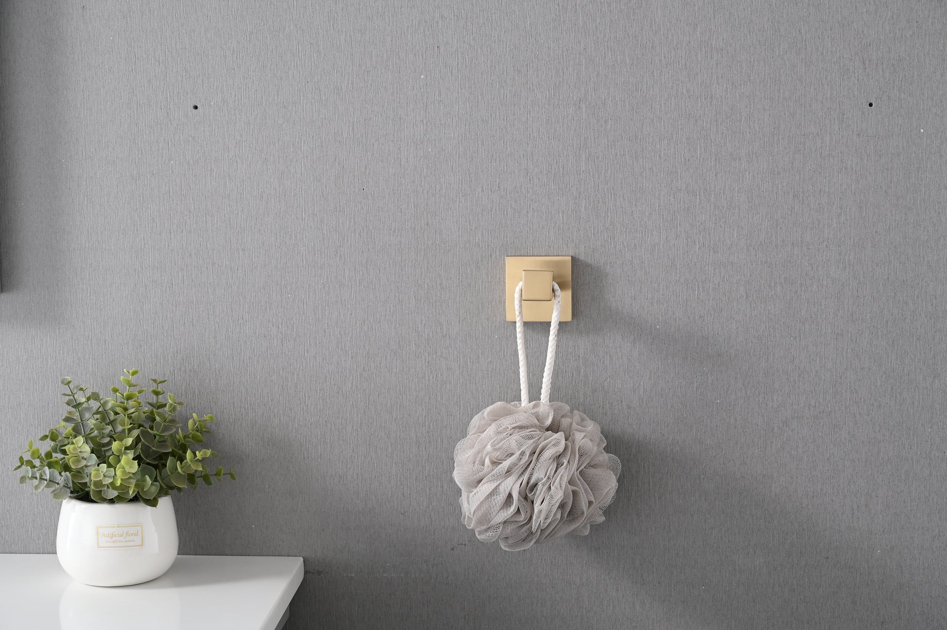 Soft beige mesh bath pouf with white rope hanging from a gold square hook