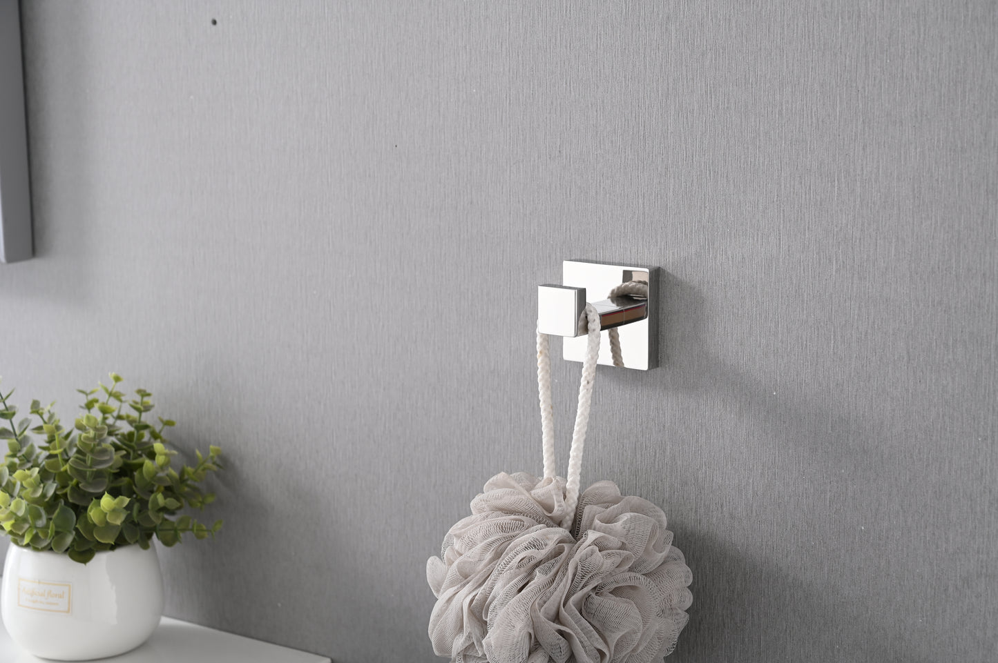 Polished chrome square hook with white rope bath pouf hanging on textured gray wall