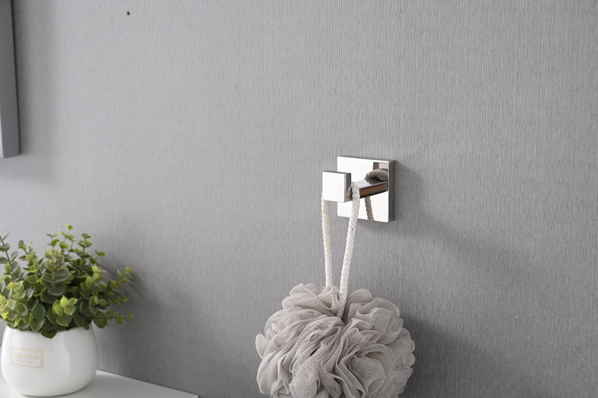 Polished chrome square hook with white rope bath pouf hanging on textured gray wall