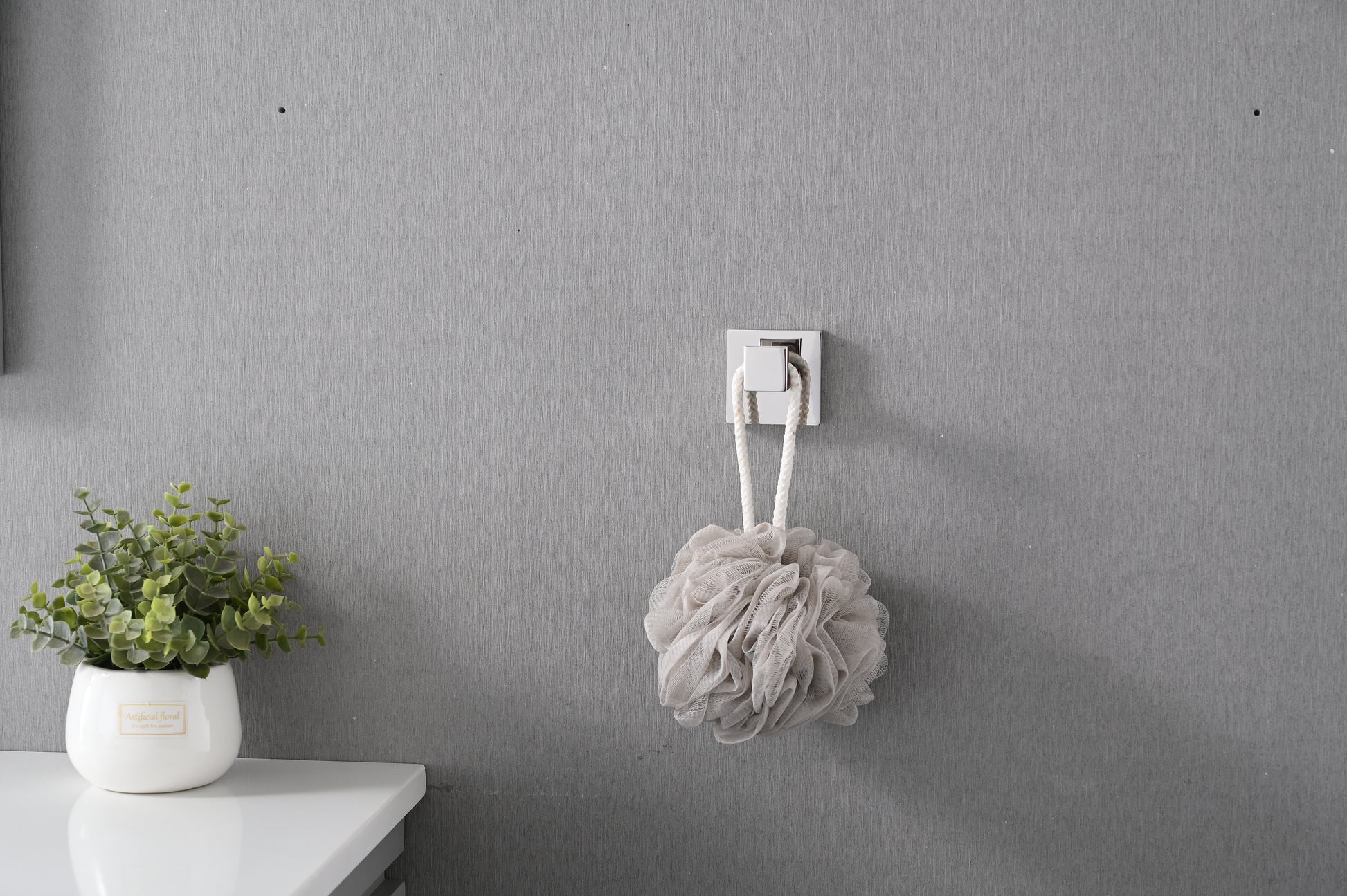Textured taupe bath pouf with white rope handle hanging on a chrome hook