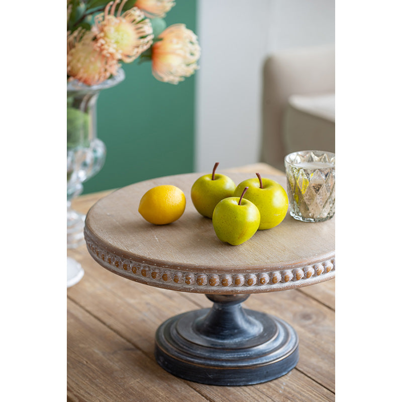 Rustic wooden cake stand with beaded rim displaying faux green apples and a lemon