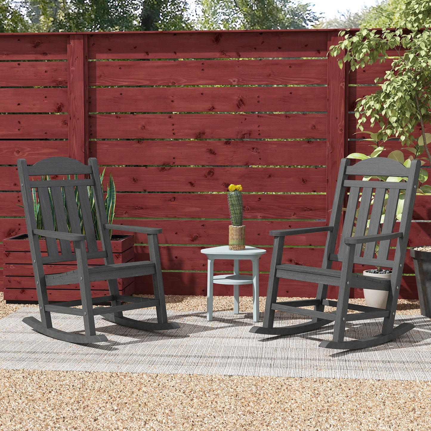 Two dark grey Adirondack rocking chairs on a patio with a red wooden fence behind
