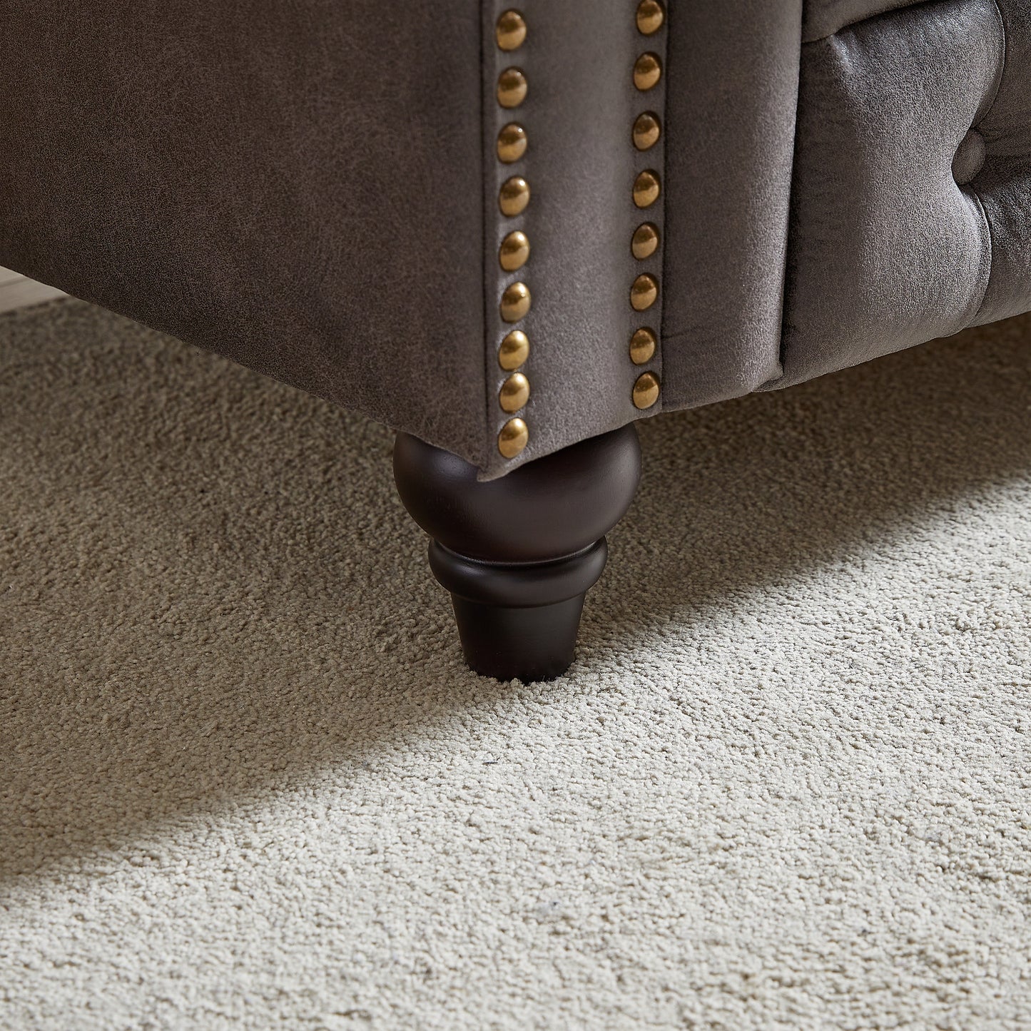 Grey microfiber sofa armrest with brass nailhead trim and dark wood leg on cream rug