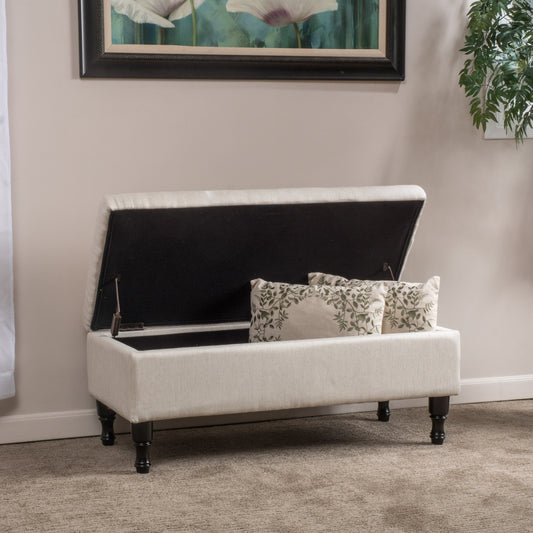Beige upholstered storage ottoman with carved dark wood legs and botanical print pillows