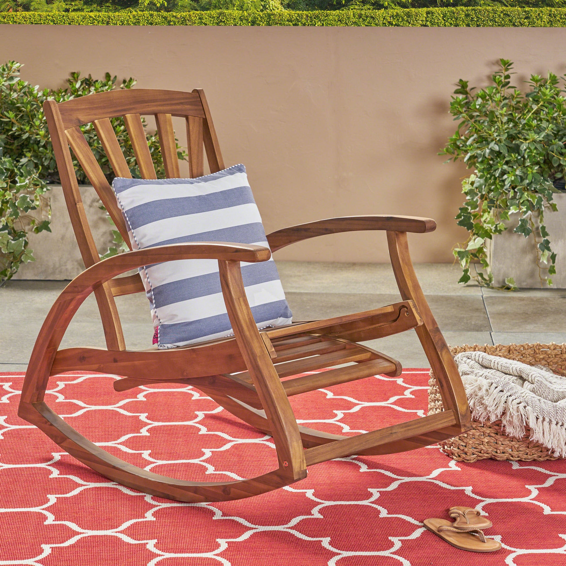 A warm wood outdoor rocking chair with a blue and white striped cushion sits on a red and white patterned rug