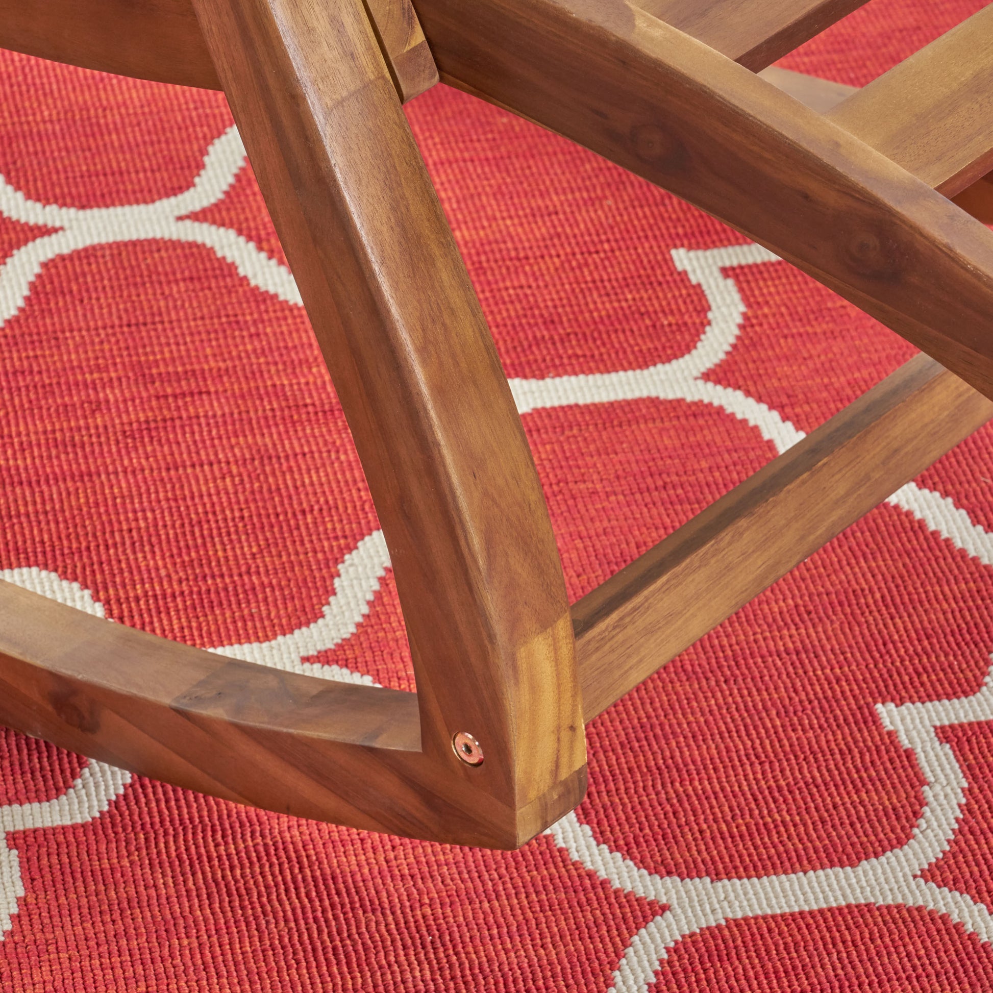 Closeup of a curved polished wooden chair leg resting on a red outdoor rug with a white geometric pattern