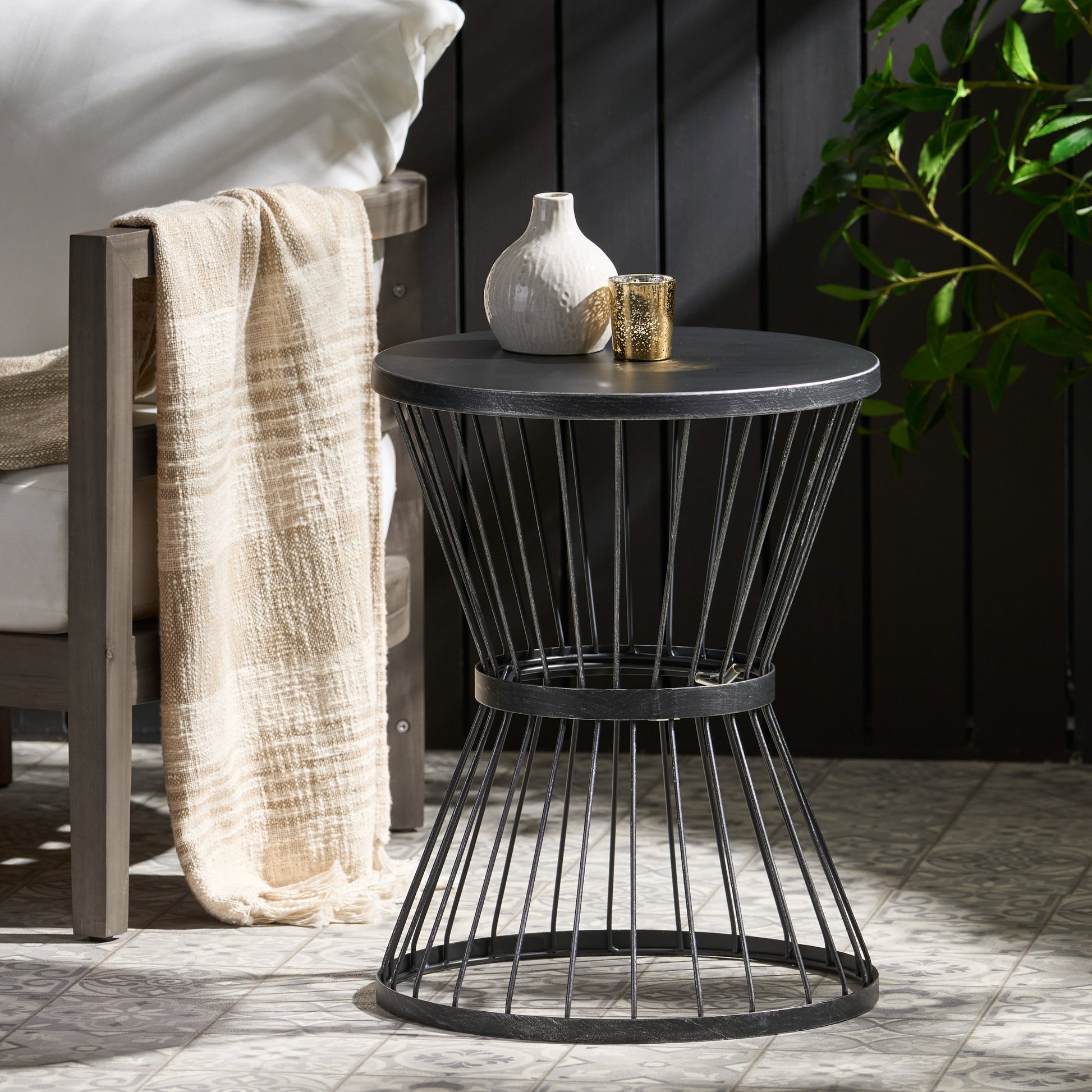 Black metal outdoor end table with spindle base and round top holding a vase and candle holder