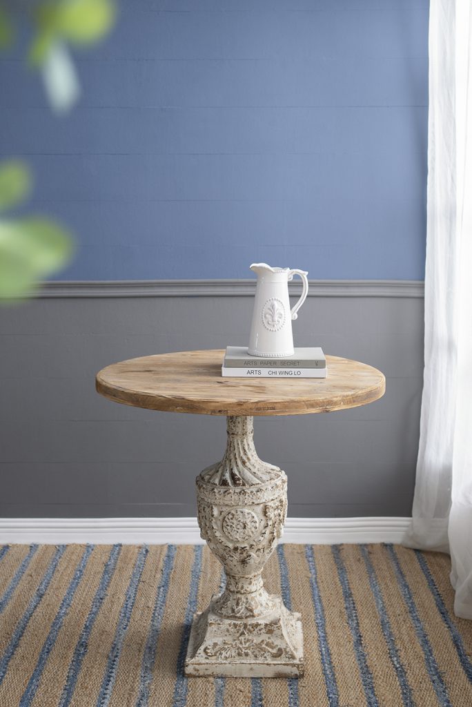 Distressed white decorative pitcher with fleurdelis motif on rustic wooden side table