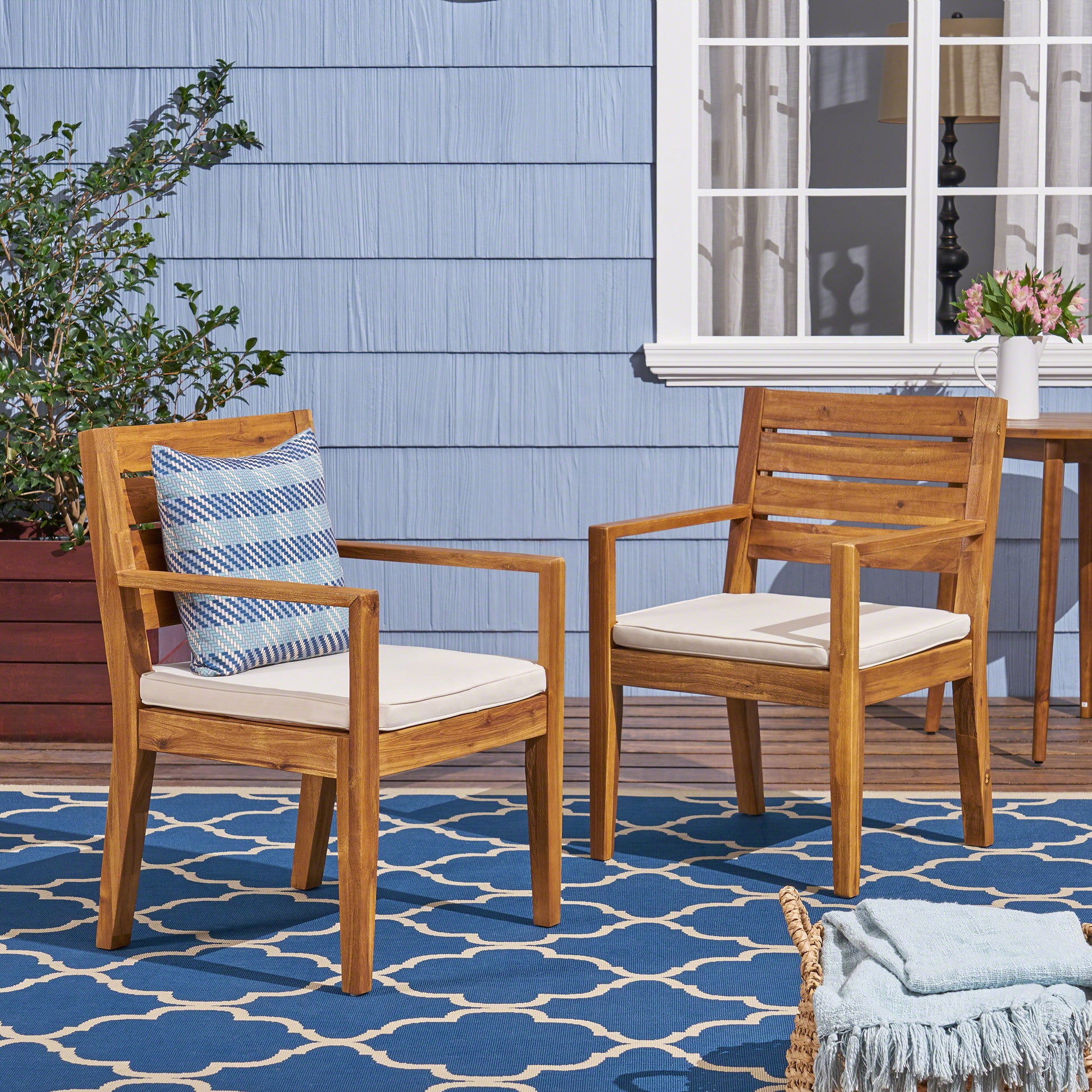Two modern acacia wood outdoor chairs with cream cushions and blue striped pillows on a patio