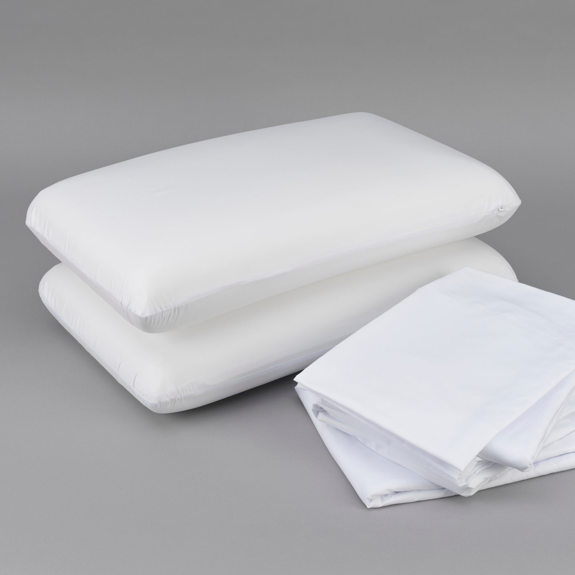 Two white down alternative pillows stacked with folded white cotton sheets on a gray background