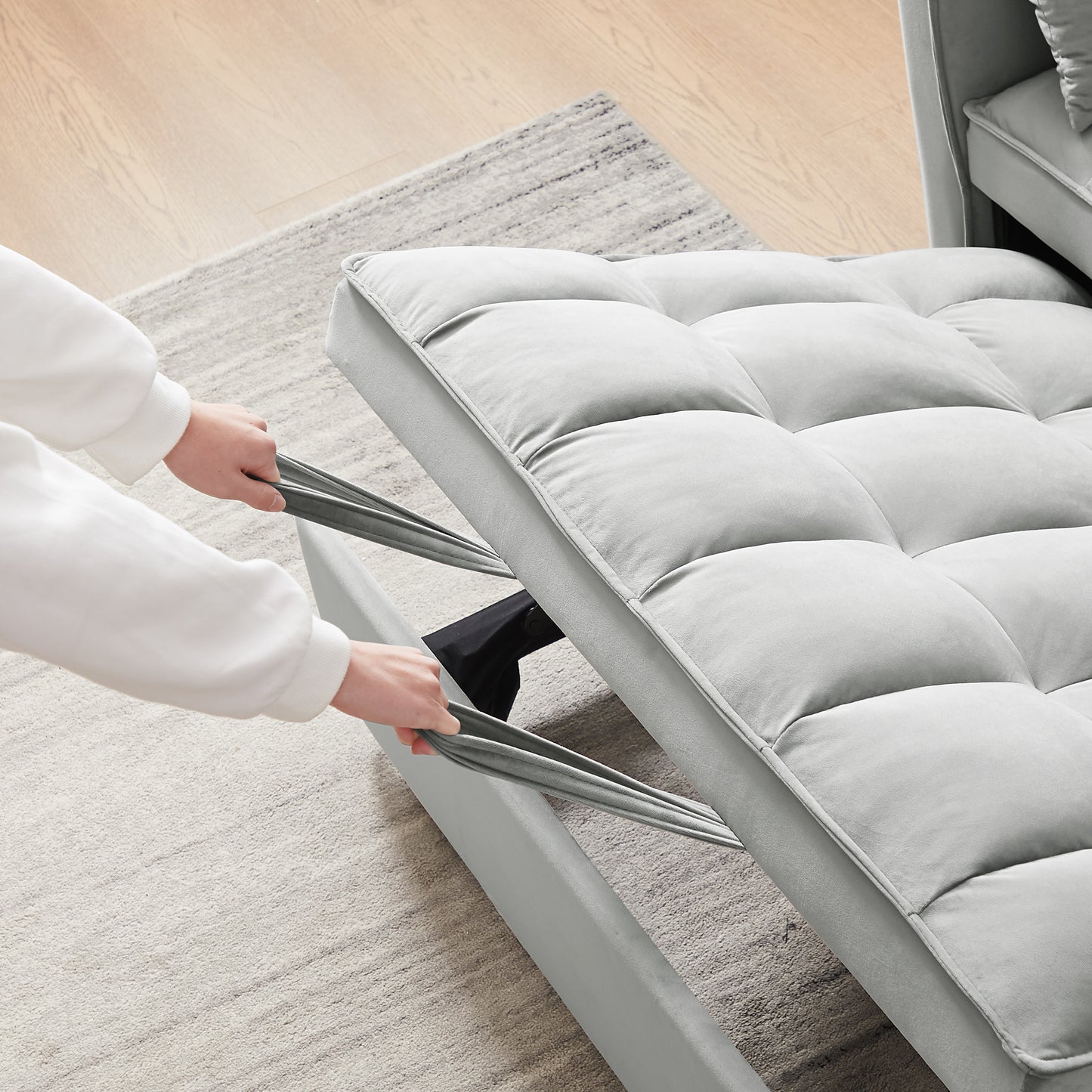 Closeup of hands pulling grey fabric straps to extend a velvet futon sofa showing tufted cushions